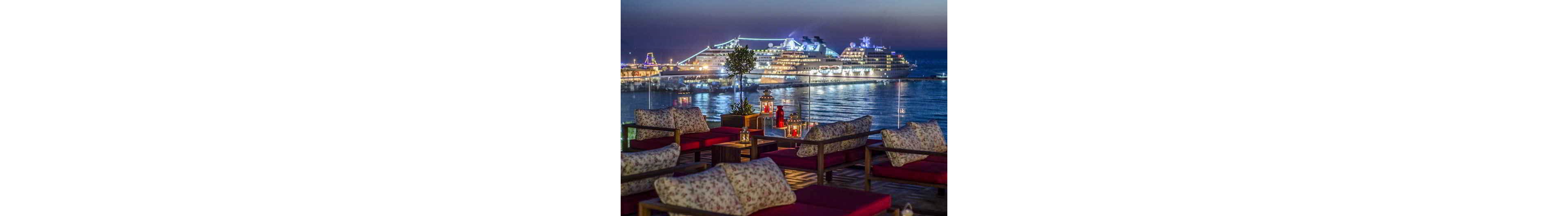 An outdoor lounge terrace with cushioned seating overlooks a brightly lit cruise ship docked in a harbor at dusk. Red cushions and floral-patterned pillows create a cozy foreground, while decorative lanterns and small tables add warmth to the scene. The large cruise ship glows with white and blue lights, reflecting on the calm water. The sky transitions from deep blue to purple, enhancing the relaxed, upscale evening atmosphere.
