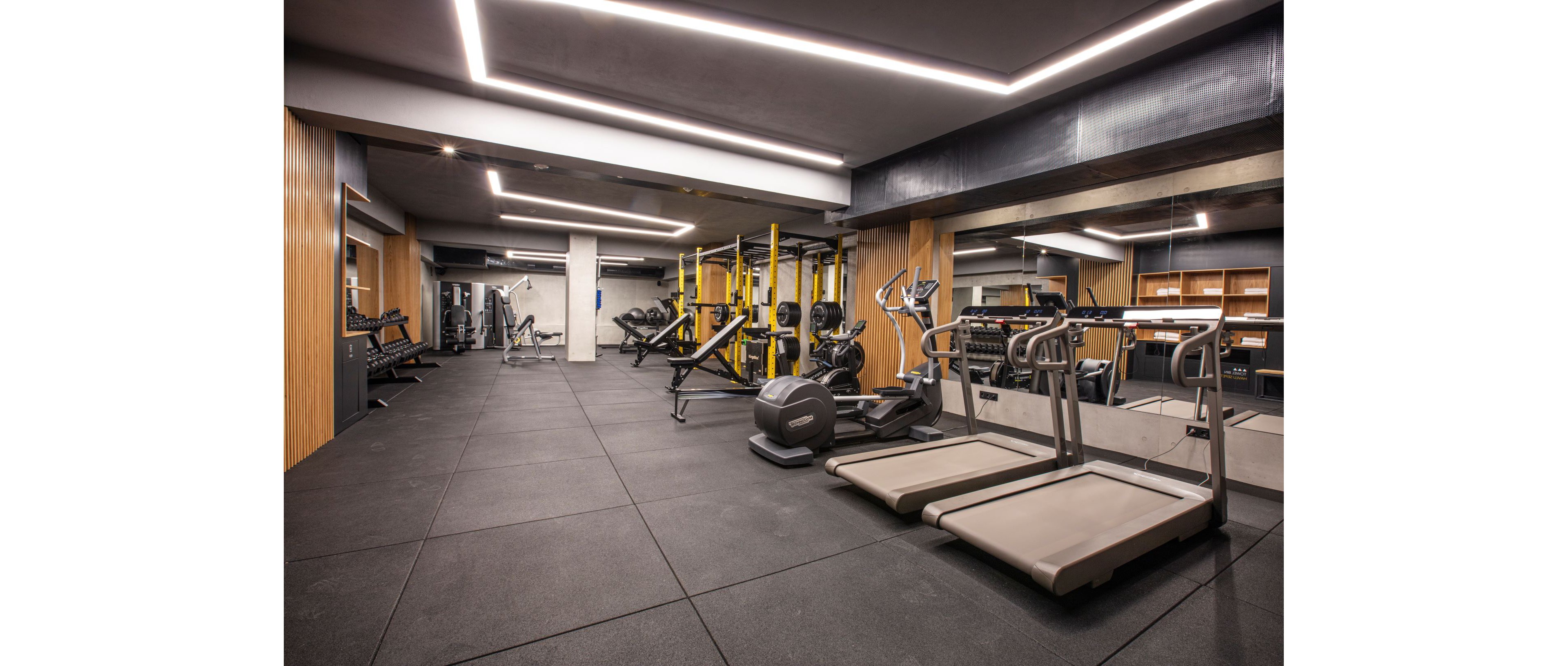 A contemporary indoor gym features a variety of fitness equipment neatly arranged in a spacious room. Treadmills and an elliptical trainer are positioned in the foreground, while weight benches, racks, and yellow functional training frames occupy the center. Large mirrors line one wall, visually expanding the space and reflecting the machines. The room has clean lines, wood panel accents, and bright linear ceiling lights, creating a sleek and professional workout environment.