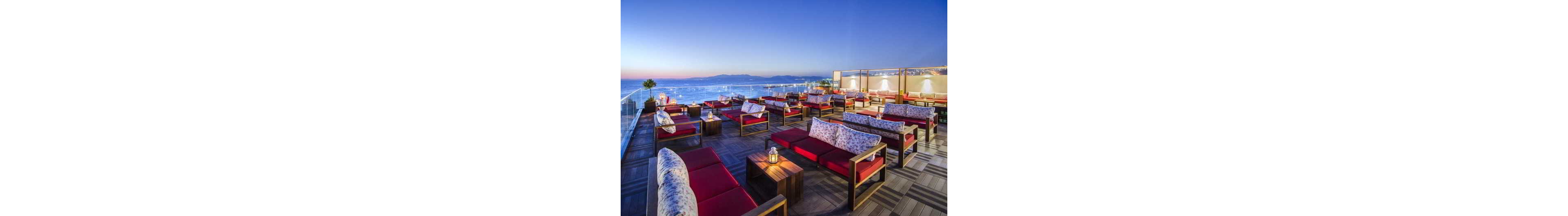 A stylish rooftop lounge features rows of red cushioned sofas and low wooden tables arranged neatly on a patterned deck. Glass railings frame a panoramic view of the ocean and distant mountains under a clear evening sky. Small lanterns on the tables create a warm, inviting glow as city lights begin to appear along the coastline. The scene suggests a relaxed, upscale outdoor setting ideal for socializing at sunset.