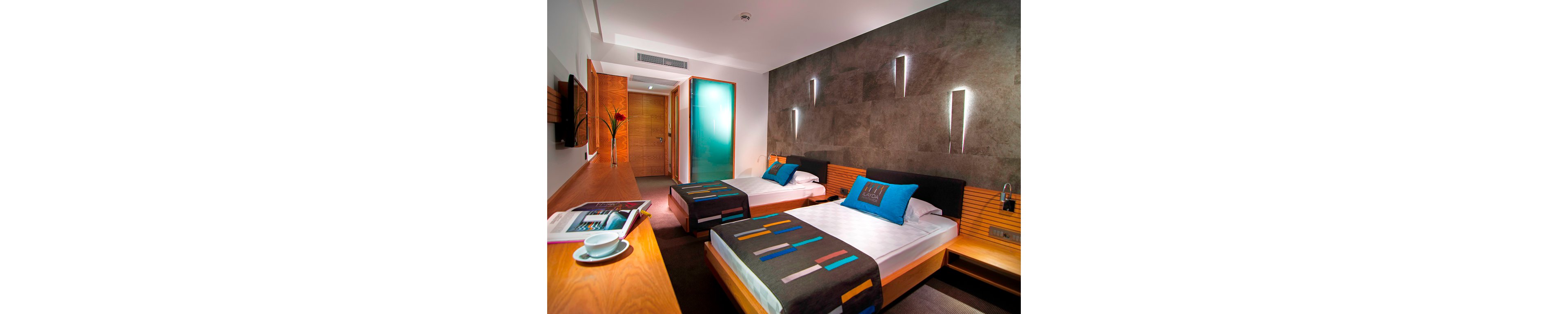 A contemporary hotel room features two neatly made twin beds with colorful geometric bed runners and bright accent pillows. The space includes a long wooden desk with an open magazine and a cup and saucer in the foreground. A frosted glass door leads to the bathroom area, while recessed lighting and wall sconces create a warm, inviting ambiance. Neutral walls and sleek furniture emphasize a modern, minimalist design.