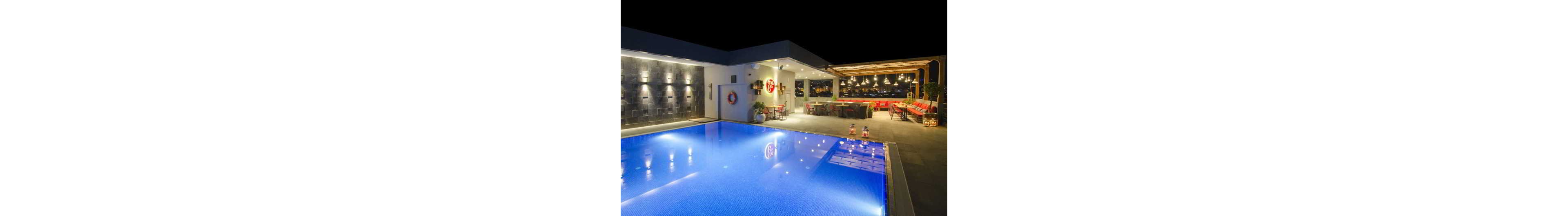 A contemporary rooftop swimming pool glows with blue lighting against the night sky. Adjacent to the pool, a stylish open-air lounge features a long bar counter and red cushioned seating. Warm pendant lights and wall sconces create a cozy, inviting atmosphere. Decorative elements like plants, lanterns, and a lifebuoy add character to the sophisticated outdoor space.