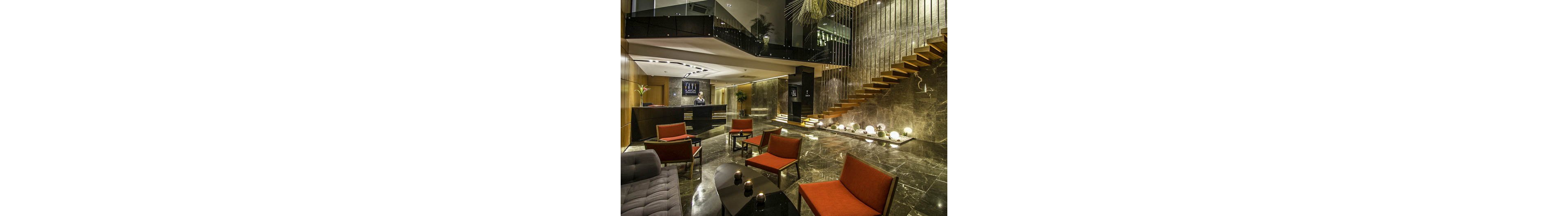 A contemporary hotel lobby features polished dark marble floors and walls with warm wood accents. Red upholstered lounge chairs and low black tables create an inviting seating area in the foreground. A reception desk with a staff member stands in the background beneath a sleek ceiling design. Floating wooden stairs and decorative spherical lights add a sophisticated, modern touch to the space.