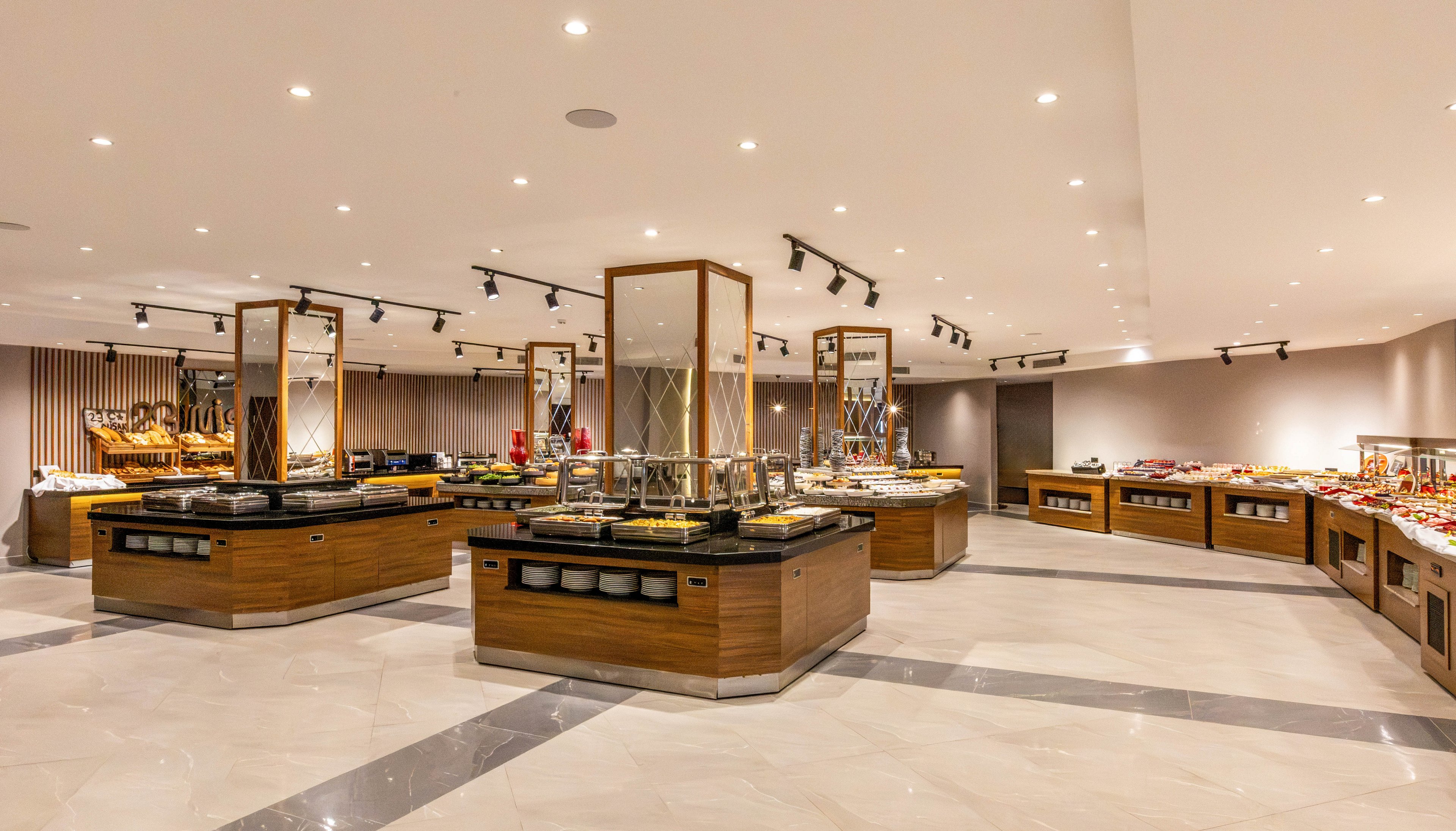 Spacious self-service buffet restaurant featuring multiple wooden food stations. The room has bright ceiling spotlights and a polished light-colored tile floor. Various dishes and serving trays are arranged neatly around the central islands and along the walls. The overall atmosphere is clean, contemporary, and prepared for guests.