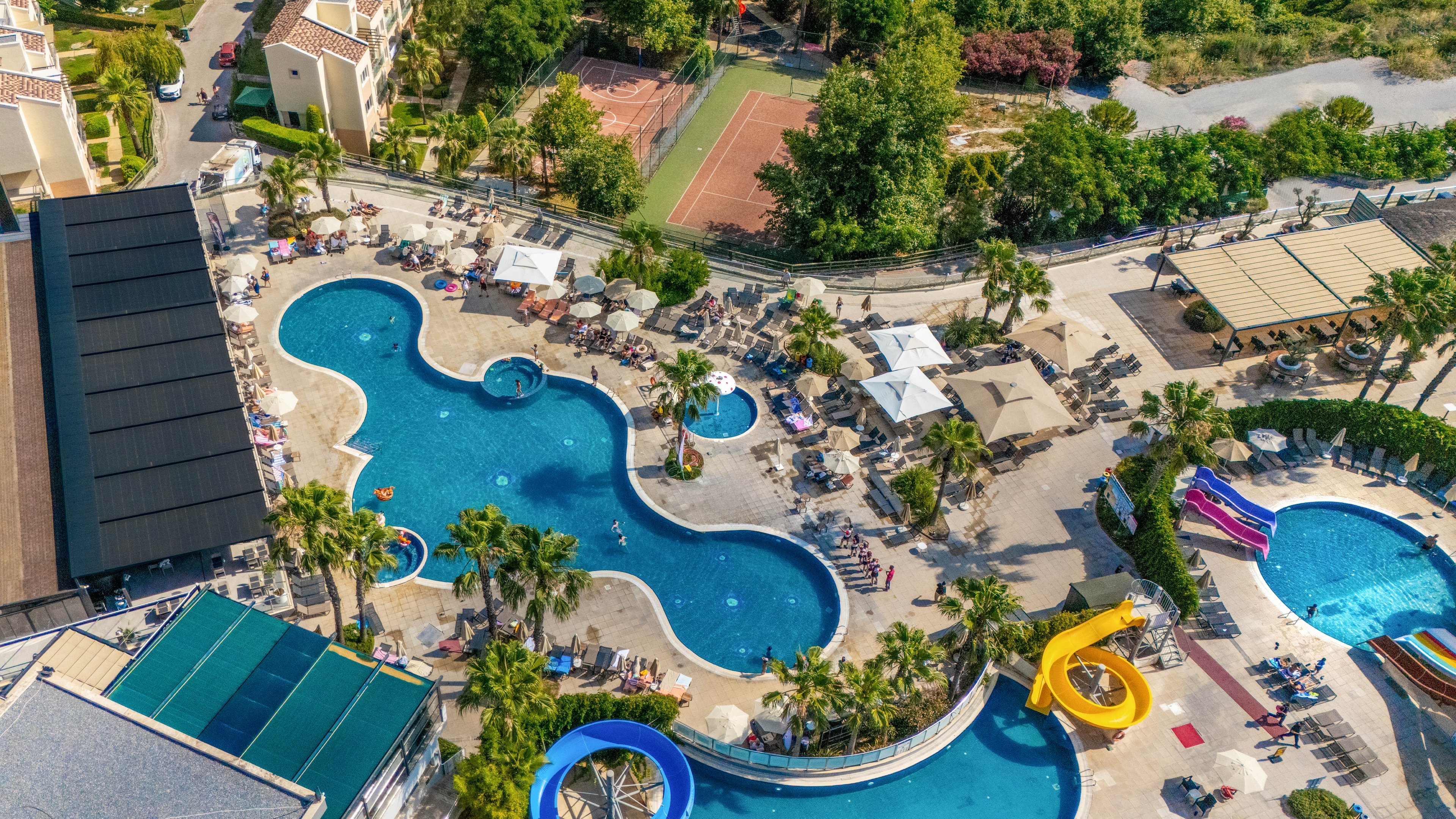 A large resort complex features multiple interconnected outdoor swimming pools surrounded by sun loungers and umbrellas. Colorful waterslides curve into a circular pool, adding a playful element to the scene. Palm trees and landscaped greenery frame the pool area, while nearby buildings and sports courts indicate a full-service vacation destination. The bright blue water and vivid slide colors create a lively, family-friendly atmosphere.