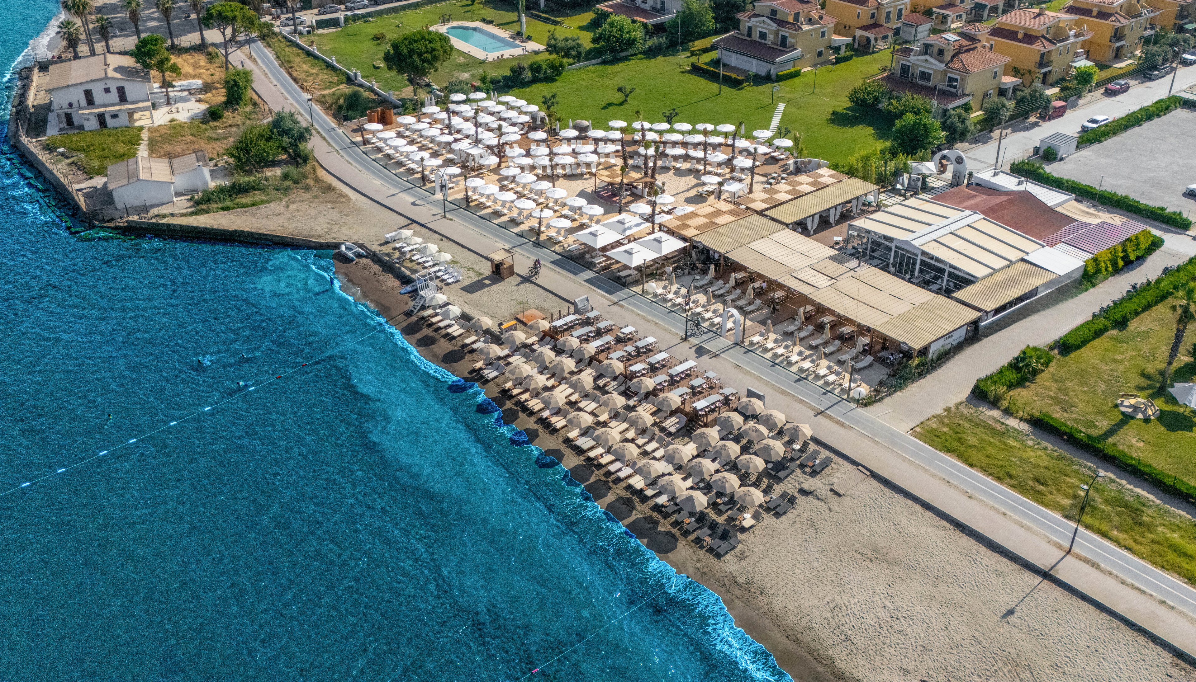 A coastal resort with rows of sun loungers and umbrellas lines a sandy beach beside vivid blue sea. The image is captured from above, showing organized beach seating areas and adjacent open-air terraces. A road separates the beachfront facilities from nearby villas, gardens, and a swimming pool. The scene suggests a sunny vacation destination with clear water and relaxed leisure spaces.