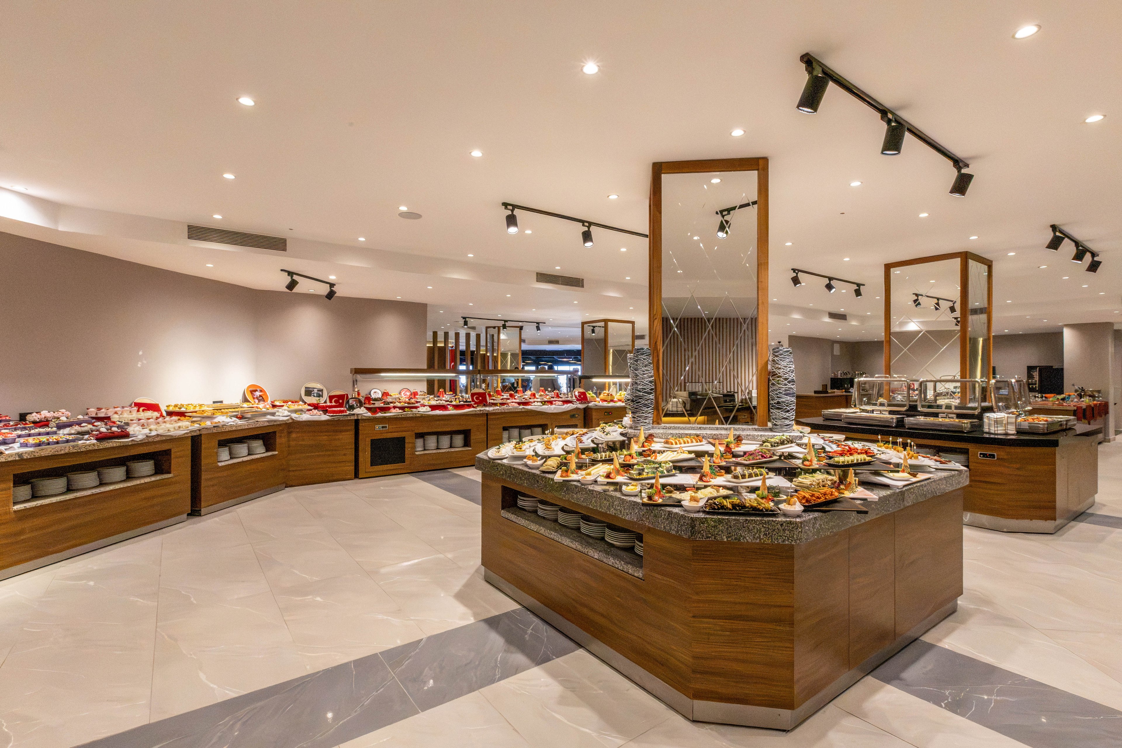 A spacious, modern buffet area showcases a wide variety of food arranged on multiple wooden islands with stone countertops. The setting appears to be an upscale hotel or restaurant dining room with bright ceiling lights and clean, polished flooring. Numerous plates, bowls, and serving trays display colorful appetizers, main courses, and desserts. Stainless steel equipment and beverage dispensers line the back counters, emphasizing a self-service dining experience.