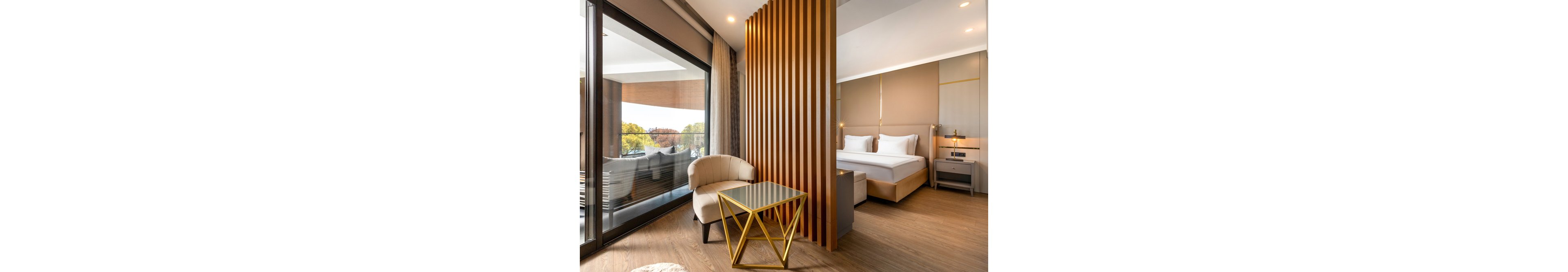 Contemporary hotel room interior featuring a comfortable bed and a stylish seating area. A geometric gold side table and upholstered armchair sit beside a vertical wooden slat divider. Floor-to-ceiling windows open to a balcony with outdoor seating and views of trees. Warm neutral tones, soft lighting, and clean lines create a cozy yet sophisticated atmosphere.