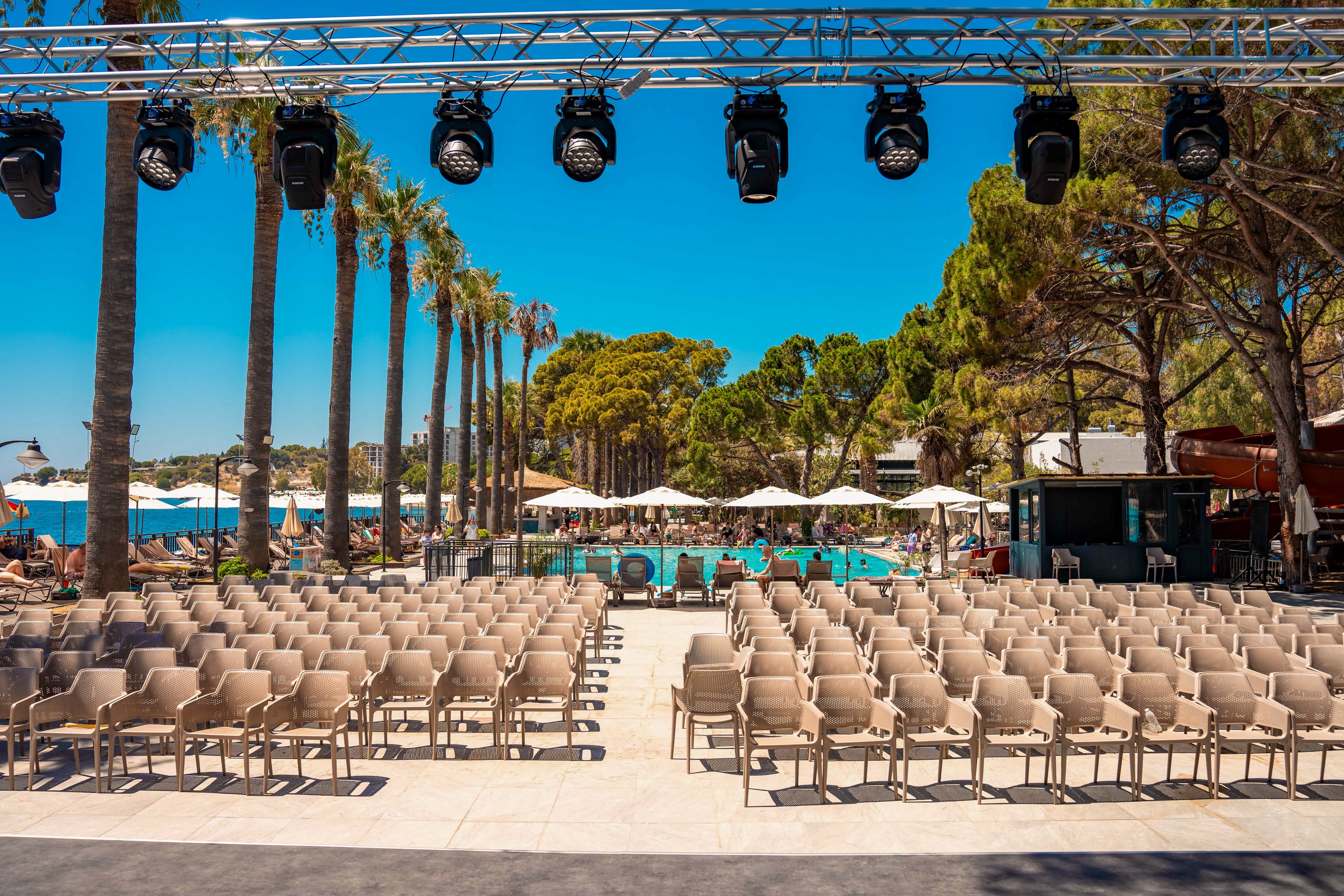An outdoor event setup featuring rows of beige chairs arranged in front of a poolside venue. The setting is surrounded by tall palm trees and lush greenery under a bright blue sky. Overhead stage lighting is visible, suggesting a performance or gathering. The scene exudes a vibrant and sunny atmosphere, ideal for a summer event.