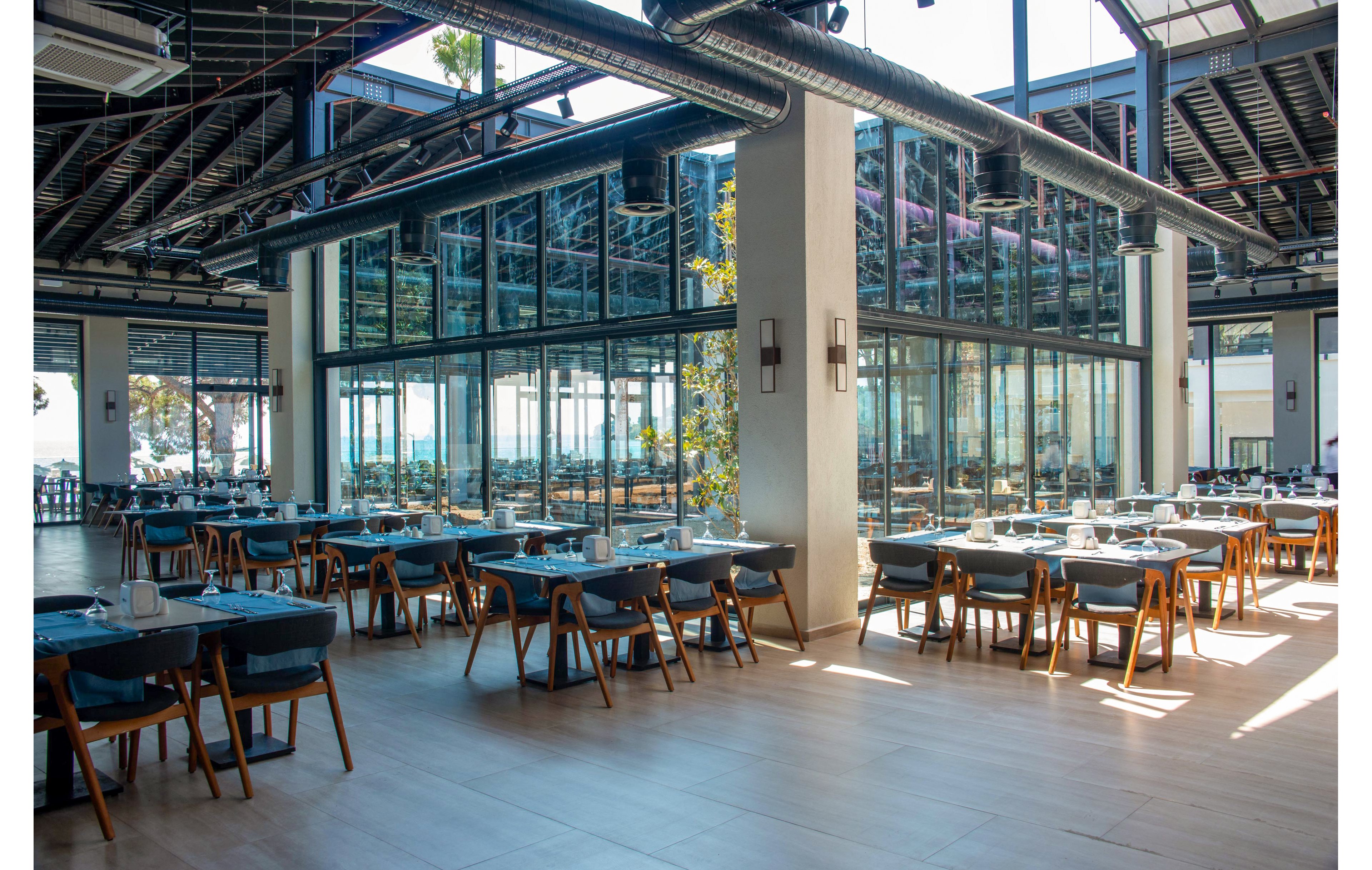 A spacious, modern restaurant featuring glass walls and wooden furniture. The setting includes neatly arranged tables with blue tablecloths and chairs, creating a clean and inviting atmosphere. Natural light streams in through large windows, enhancing the open and airy feel of the space.