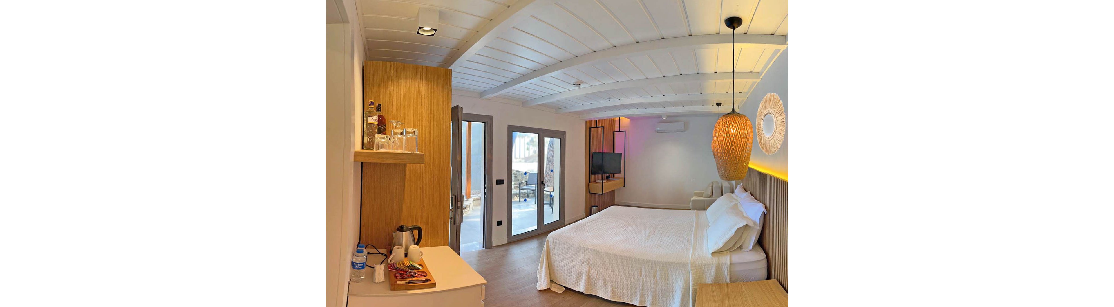 A bright and modern hotel room featuring a neatly made bed with white linens, wooden accents, and soft ambient lighting. The setting includes a tea kettle, bottled water, and a minibar on a counter. Large glass doors open to an outdoor view, enhancing the airy atmosphere.