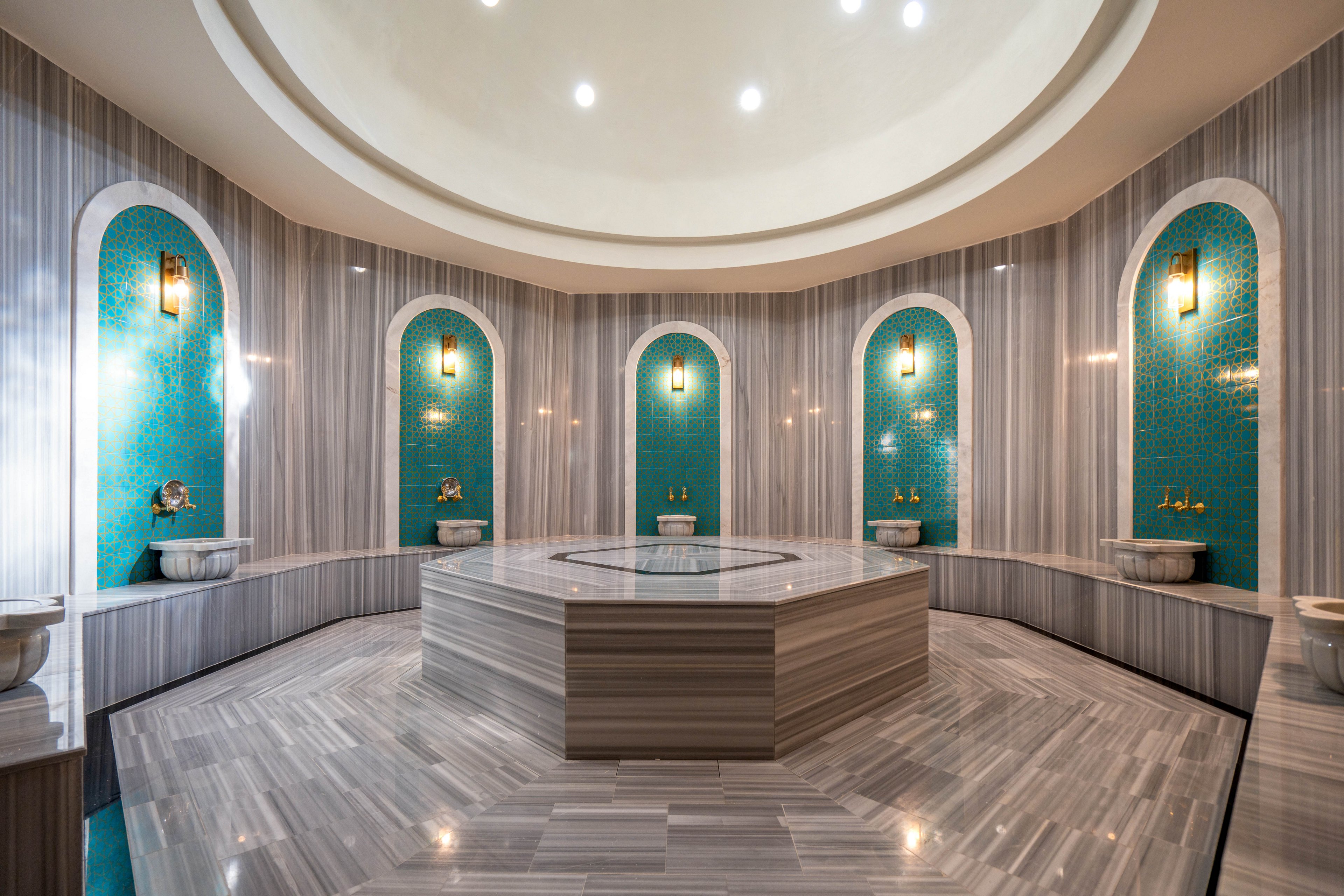 A spacious Turkish bath featuring a central hexagonal marble platform surrounded by teal mosaic walls and arched niches. The setting is illuminated by warm wall-mounted lights and a domed ceiling with recessed lighting. The elegant design combines modern marble textures with traditional Turkish aesthetics.