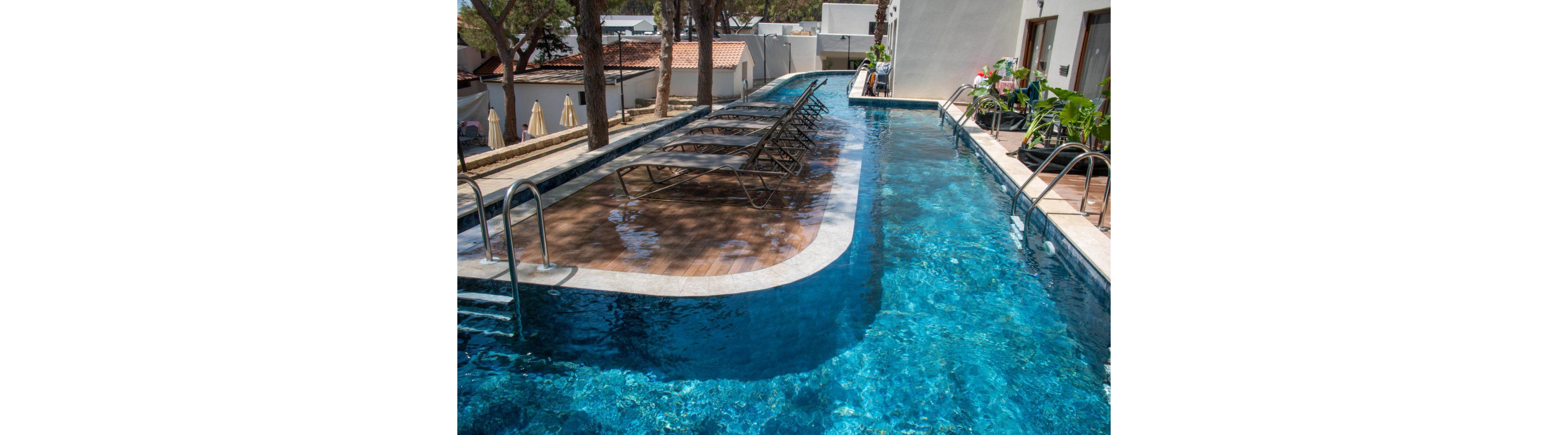 A sleek outdoor swimming pool surrounded by lounge chairs on a wooden deck. The setting is bright and sunny, with trees and residential buildings visible in the background. The pool features clear blue water and a contemporary design, creating a relaxing atmosphere.