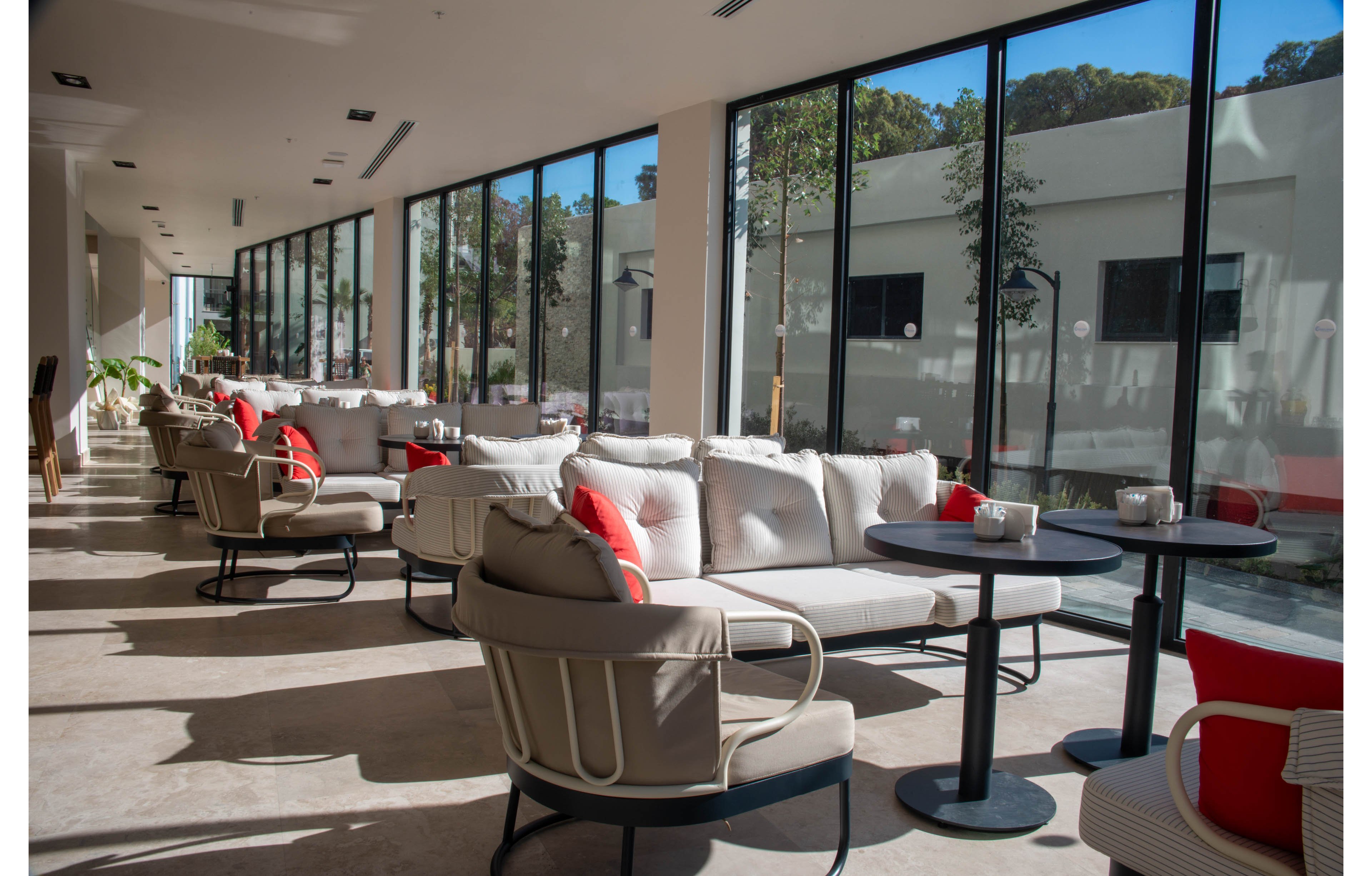 A spacious and modern lounge area featuring comfortable seating with neutral tones and red accents. Large floor-to-ceiling windows allow ample natural light to fill the space, offering views of greenery outside. The setting is ideal for relaxation or casual meetings, with tables and chairs arranged for convenience.