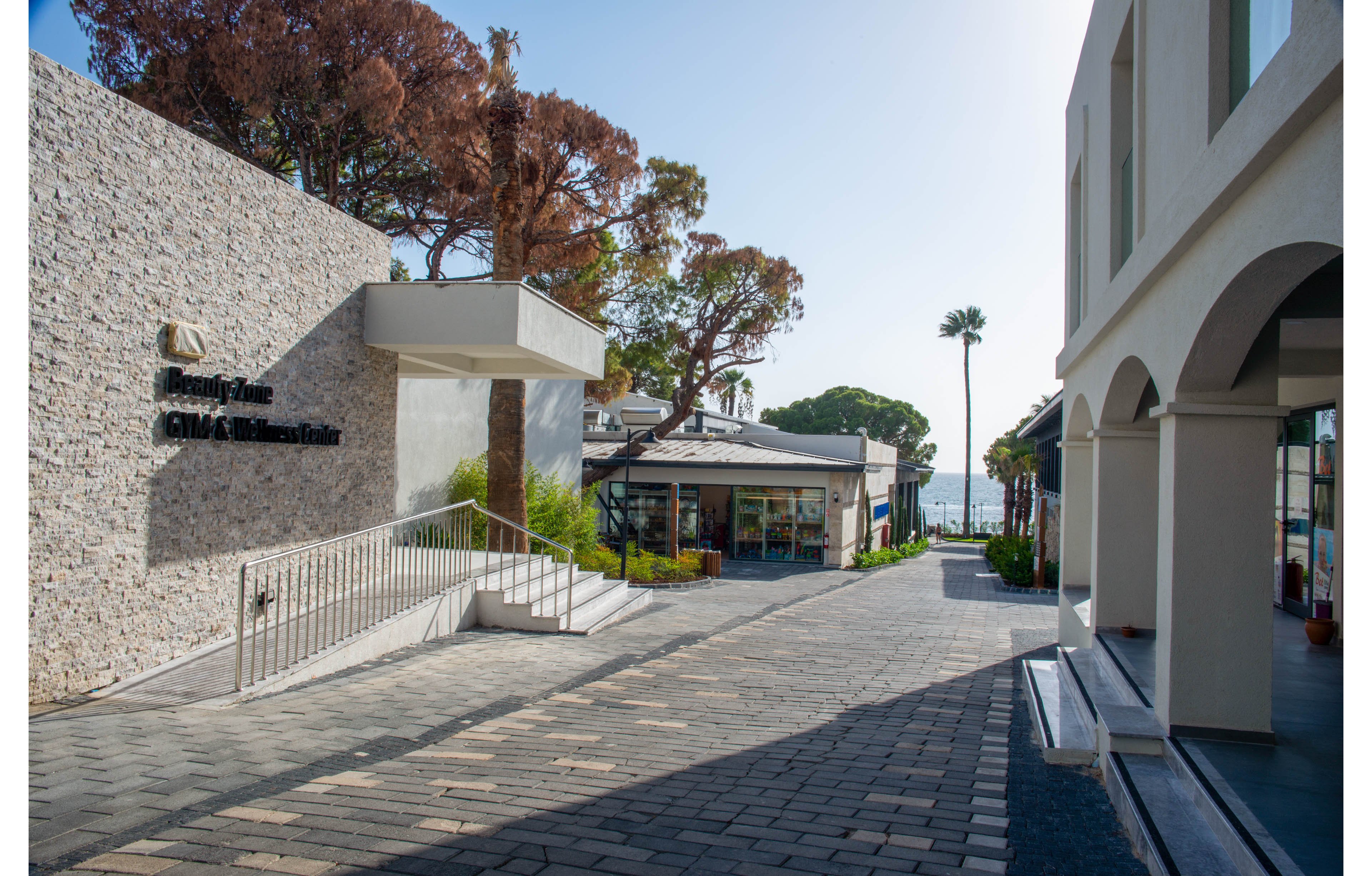 A contemporary shopping center with stone and white architectural elements, featuring a clear pathway leading to shops and a view of the ocean. The setting includes palm trees and greenery, creating a relaxed coastal vibe. Visible text reads 'Malaga Cove Civic Center'.