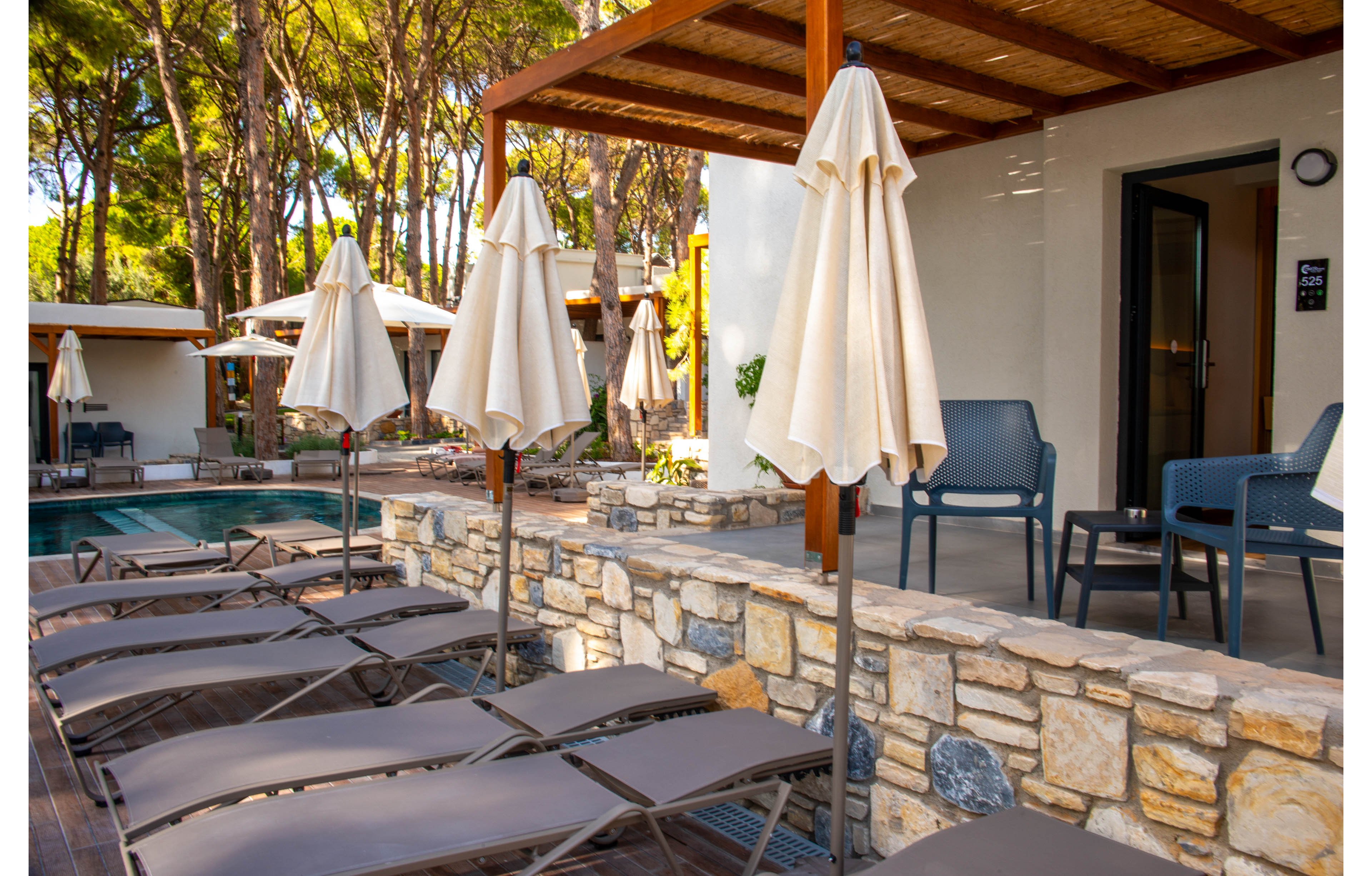 A serene outdoor poolside area featuring multiple lounge chairs and beige umbrellas. The setting includes stone walls and wooden pergolas, surrounded by lush trees. The tranquil atmosphere suggests relaxation and leisure in a natural environment.