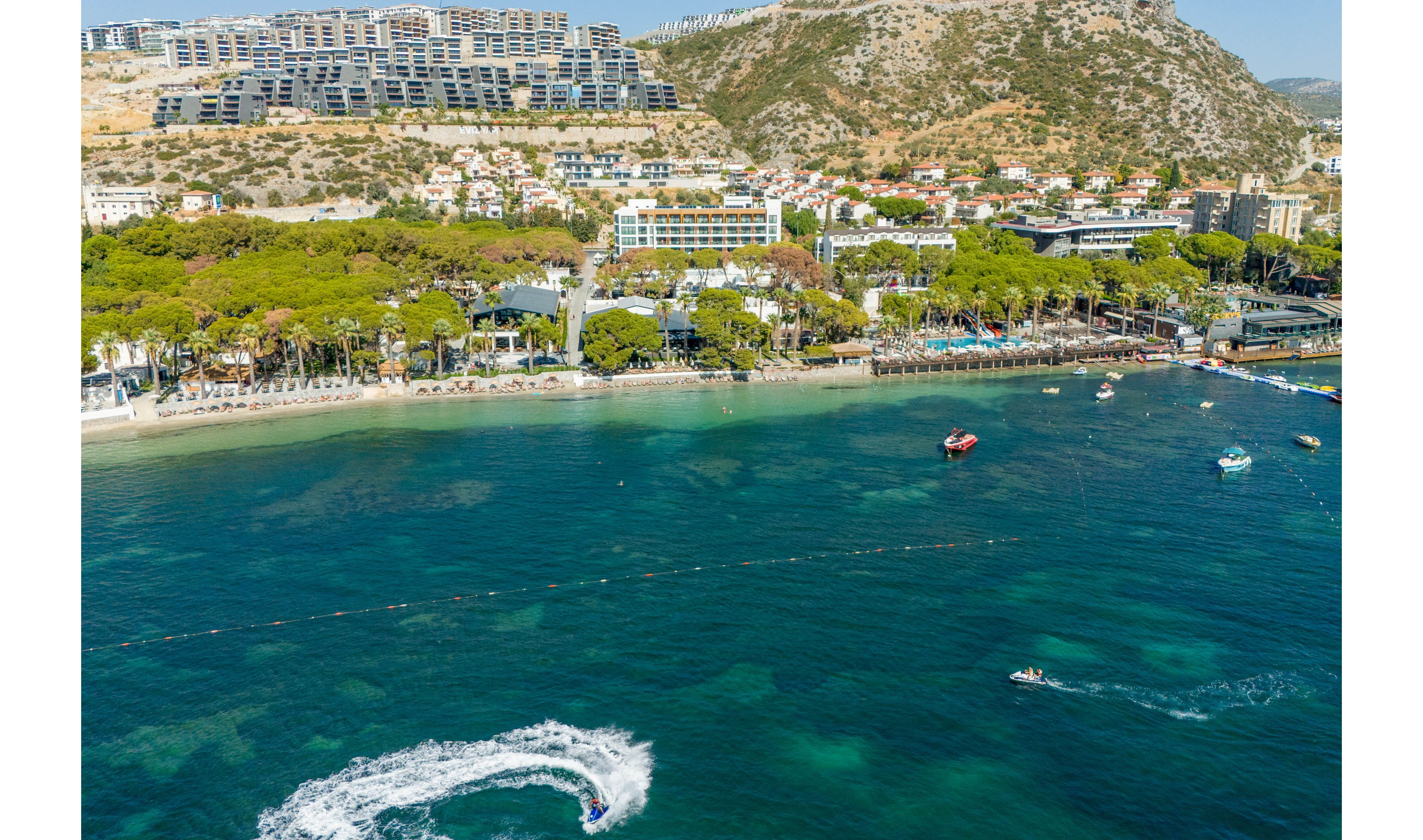 A vibrant coastal town featuring a mix of residential and commercial buildings nestled against a hillside. The foreground showcases clear turquoise waters with visible water sports activities, including jet skiing. The area is surrounded by lush greenery and a bustling beachfront, creating a lively and picturesque setting.
