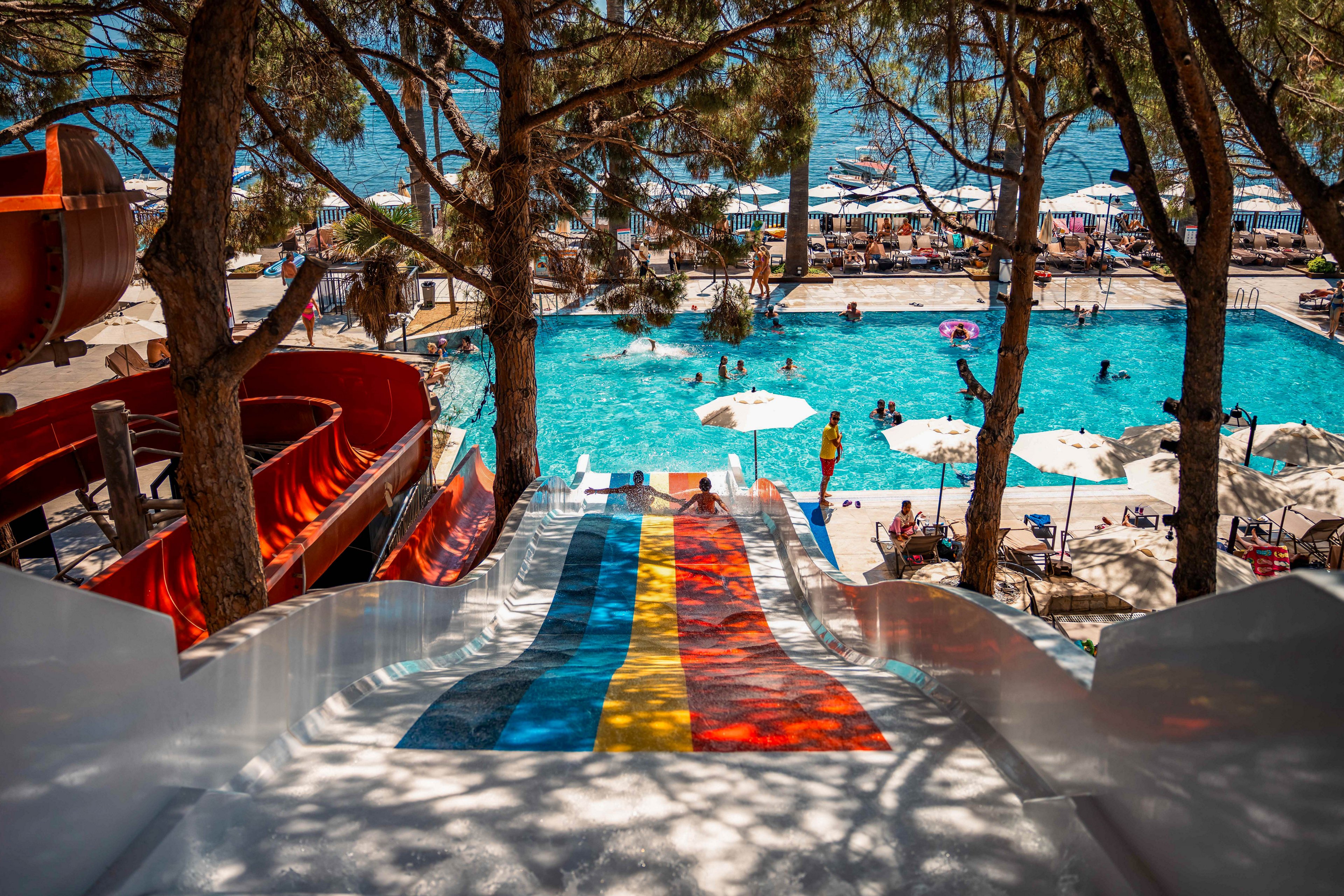 A vibrant water park featuring colorful slides in red, yellow, and blue leading into a large swimming pool surrounded by umbrellas and lounge chairs. The setting is outdoors with tall trees providing shade and a view of the ocean in the background. Families and individuals are enjoying the sunny day by swimming and relaxing.