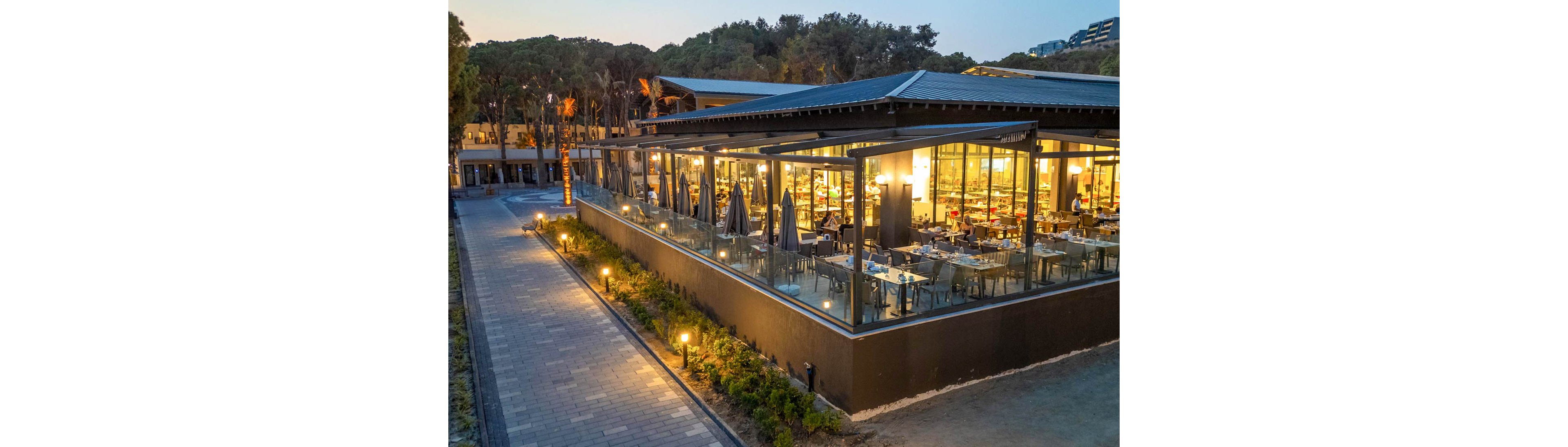 A contemporary restaurant with large glass windows and outdoor seating is illuminated during twilight. The setting features a paved walkway surrounded by greenery and trees, creating a serene atmosphere. Warm lighting enhances the inviting ambiance of the dining area.