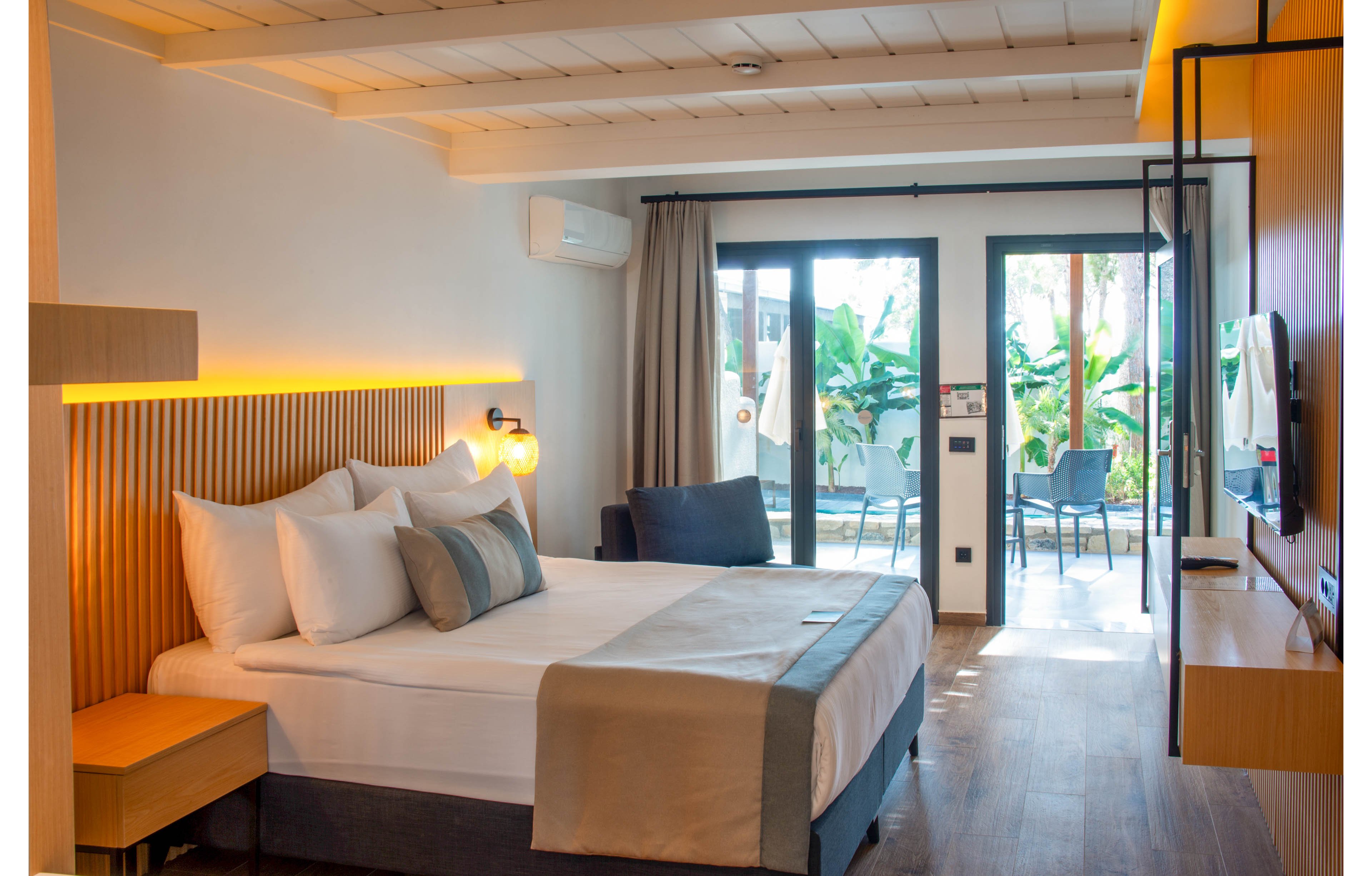 A contemporary hotel room featuring a neatly made bed with white and gray linens, wooden accents, and warm lighting. The room opens to a balcony with outdoor seating and lush greenery visible in the background. The space is bright and inviting, with a minimalist design and natural elements.