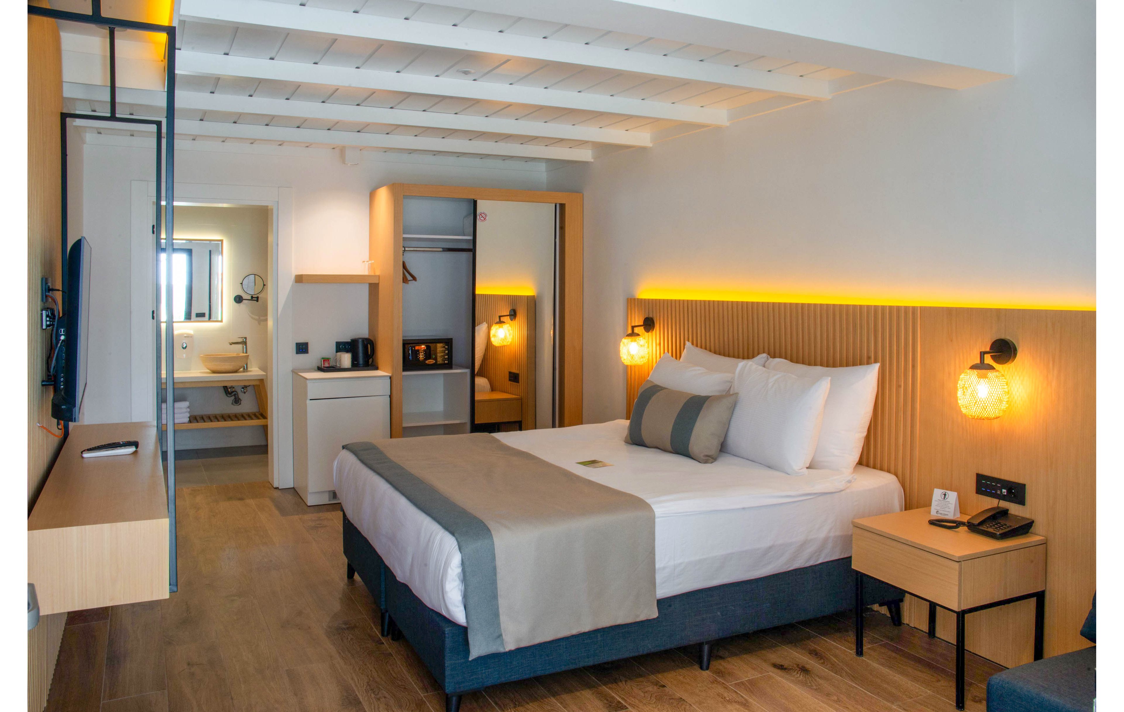 A contemporary hotel room featuring a neatly made double bed with white linens and beige accents. The room includes a small kitchenette with a microwave and mini-fridge, and a well-lit bathroom visible in the background. Warm lighting and wooden flooring create a welcoming and relaxing atmosphere.