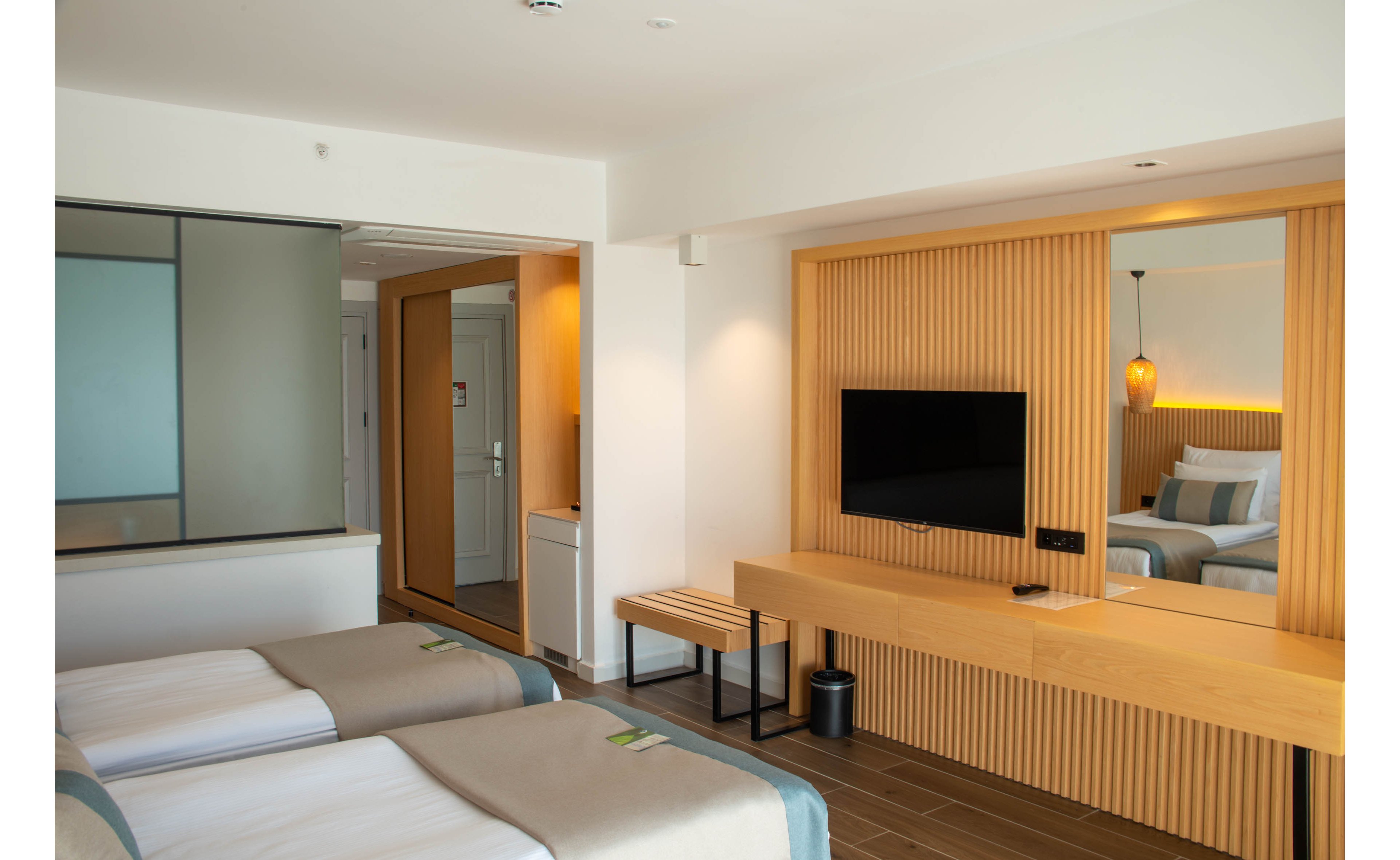 A contemporary hotel room featuring two neatly made twin beds with beige and white bedding. The setting includes a wooden accent wall, a mounted flat-screen TV, and a minimalist desk. Warm lighting and clean lines create a cozy and inviting atmosphere.