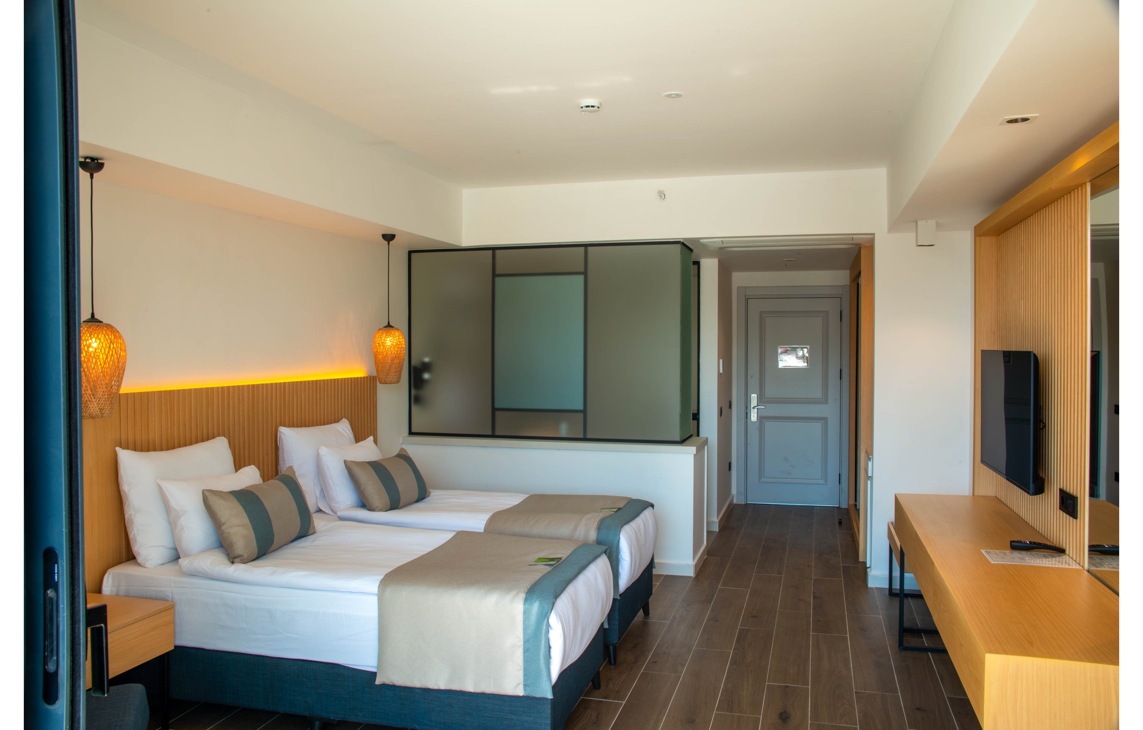 A contemporary hotel room featuring two neatly arranged twin beds with neutral-toned bedding and cushions. The setting includes wooden flooring, a desk with a mounted TV, and warm pendant lighting. The room has a minimalist design with a partition separating the sleeping area from the entrance.