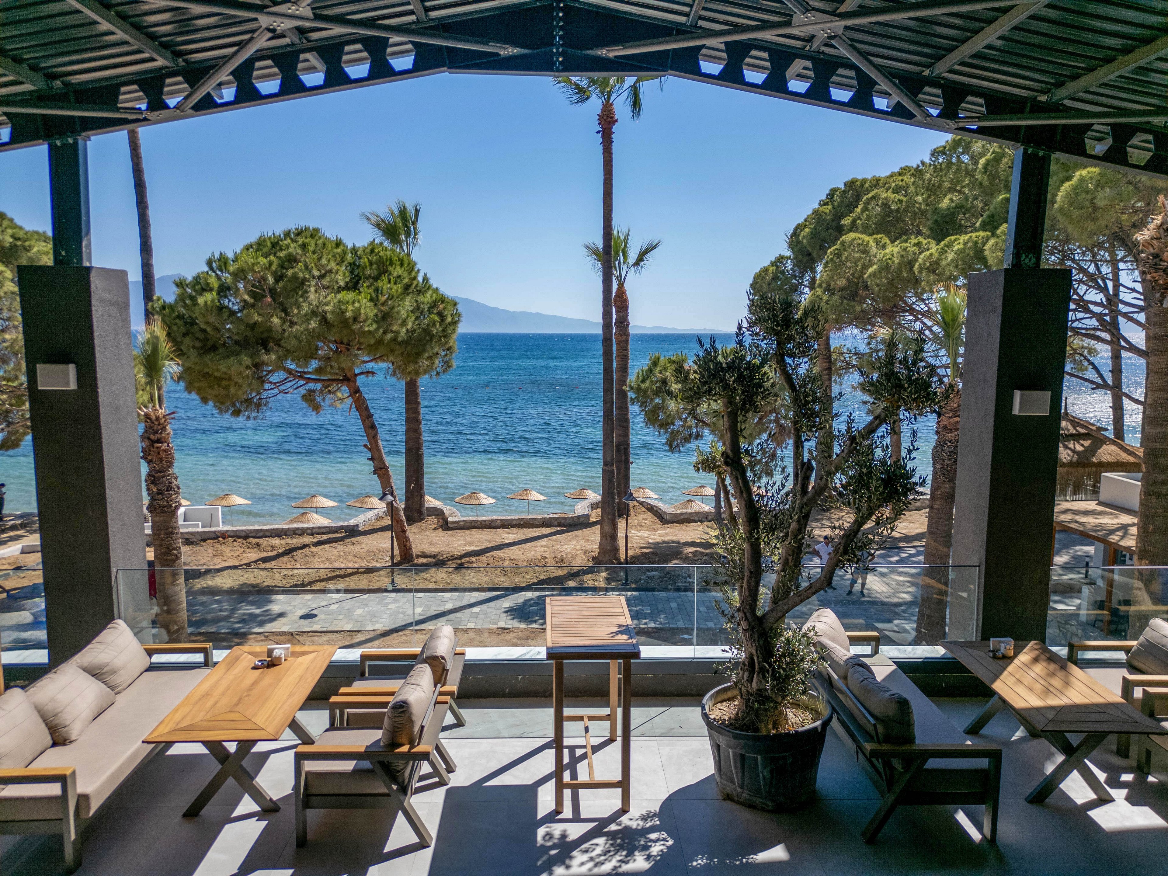 A contemporary open-air terrace with cushioned seating and wooden tables overlooks a tranquil beachfront. Sun loungers and umbrellas line the sandy shore, leading to clear blue water and distant mountains on the horizon. Tall palm trees and other greenery frame the view, while a glass railing separates the terrace from the beach. Bright daylight and clear skies create a relaxed, vacation atmosphere.