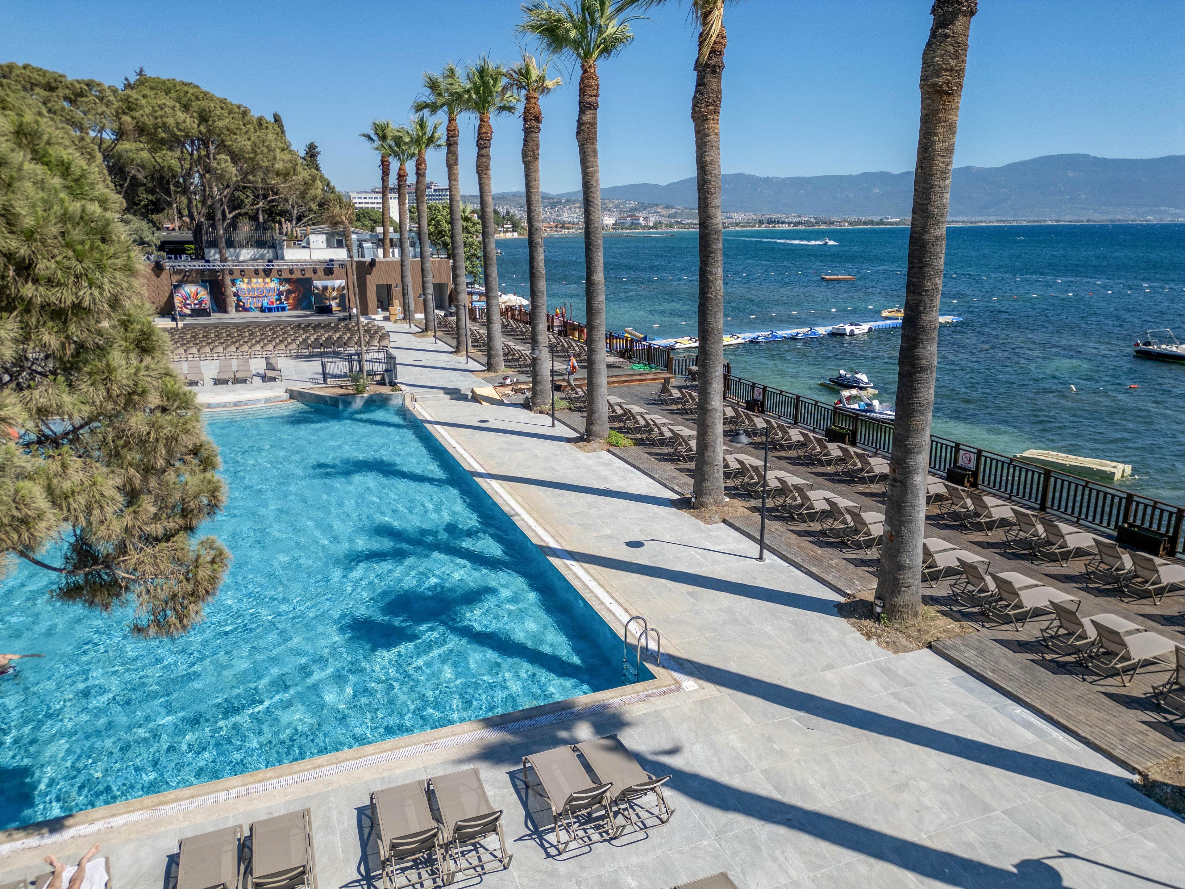 A large outdoor swimming pool sits beside a calm blue sea at a coastal resort. Tall palm trees line the pool deck, casting shadows over rows of empty lounge chairs. Guests relax near the water and on an elevated terrace overlooking the pool. The clear sky and distant mountains create a bright, tranquil vacation atmosphere.