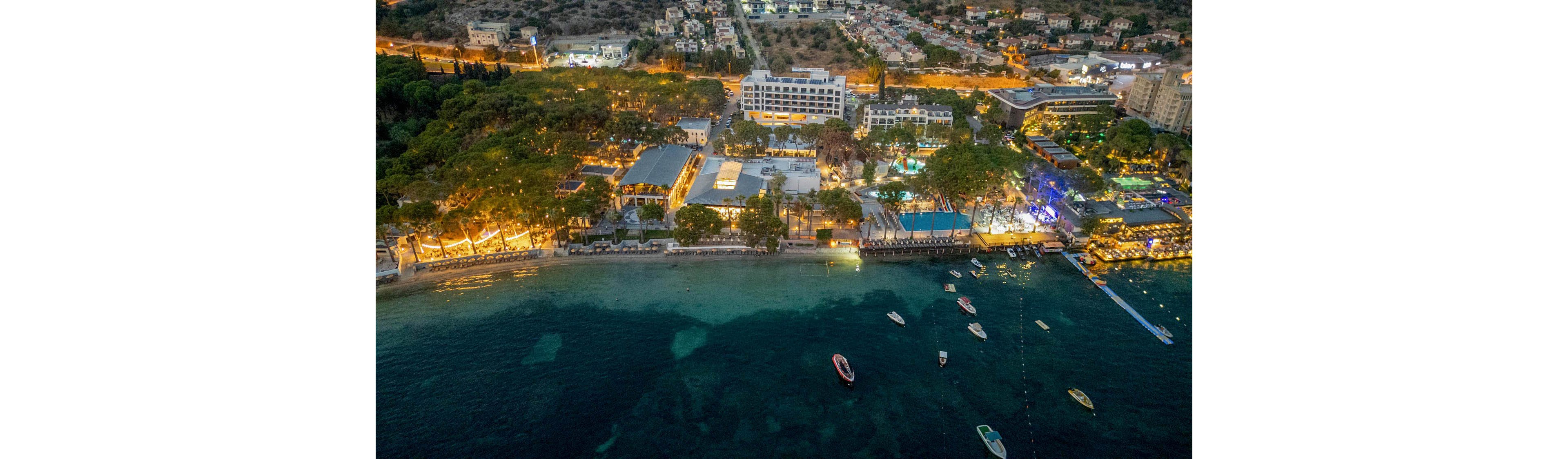 A coastal resort is captured from above at dusk, with buildings and pools brightly illuminated along the shoreline. Small boats float on the calm, shallow water near the beach, and a pier extends into the sea. The surrounding area shows dense trees transitioning into a residential neighborhood in the background. Warm artificial lights contrast with the cool blue tones of the water, creating a vibrant evening atmosphere.