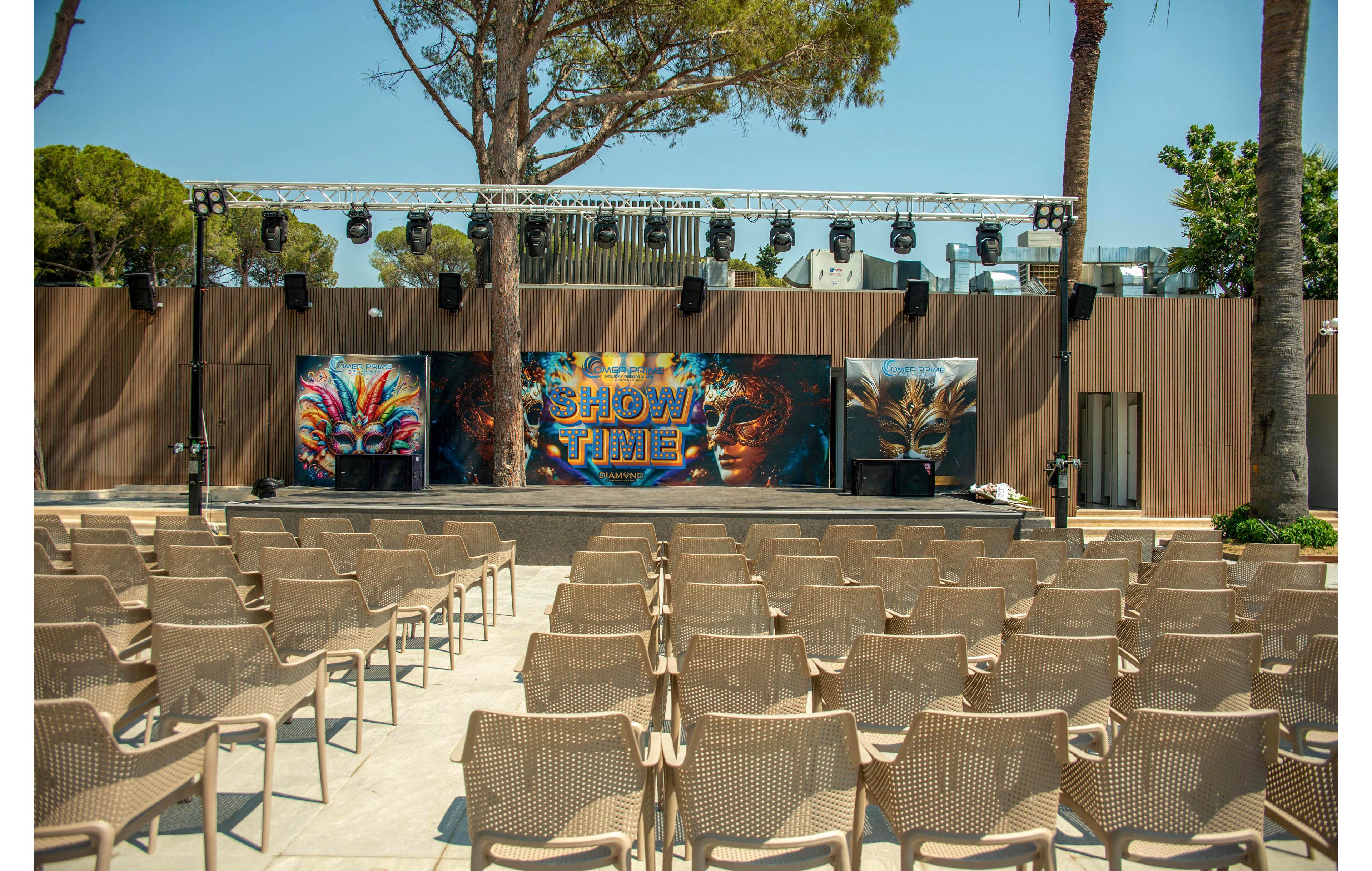 An outdoor performance stage is set up in front of rows of empty beige chairs. The backdrop features colorful carnival-style masks and a central panel with the words "SHOW TIME" in bold letters. Stage lighting equipment is mounted on a truss above the platform, ready for an event. Tall trees and a modern wooden structure frame the open-air venue under bright daylight.