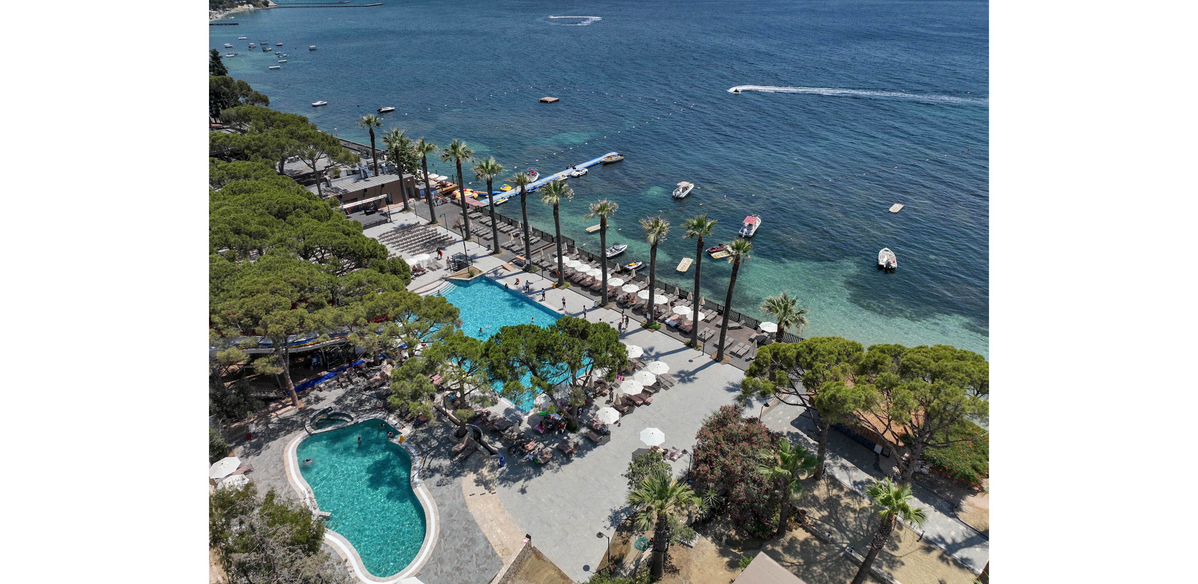 Aerial view of a seaside resort features two swimming pools surrounded by palm trees and sun loungers. The larger pool sits near a row of umbrellas and a terrace area, while a smaller curved pool is nestled among greenery. Beyond the resort, calm blue water stretches out with several small boats and watercraft visible. The scene suggests a relaxing vacation atmosphere with clear weather and coastal scenery.