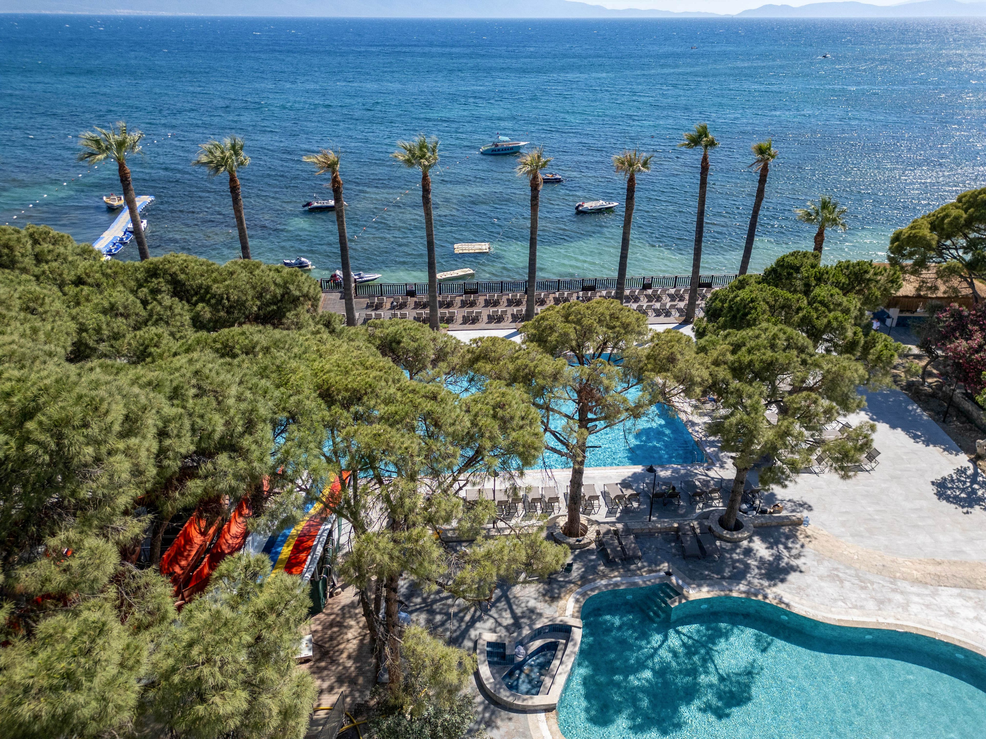 A seaside resort features multiple outdoor swimming pools surrounded by tall pine trees. Sun loungers line the beachfront, facing the calm blue water and anchored boats offshore. Bright waterslides add a splash of color near the pool area, suggesting family-friendly recreation. The overall scene conveys a sunny, relaxing holiday atmosphere on the coast.