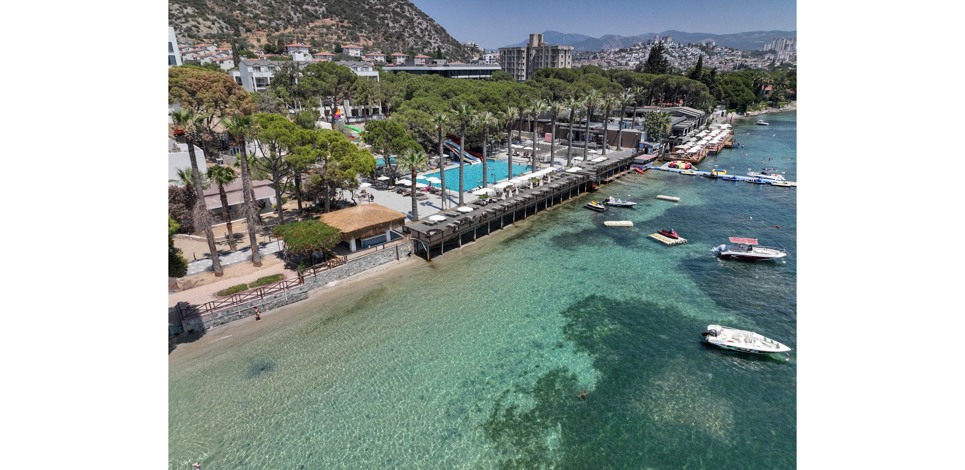An aerial view shows a seaside resort with a long waterfront lined by tall trees and sun loungers beside a large outdoor swimming pool. The complex sits along a clear turquoise shoreline where shallow water transitions into deeper blue. Several small boats and floating platforms are scattered near the coast, adding to the recreational atmosphere. In the background, hillside buildings and distant city structures rise above the bay, framing the coastal landscape.