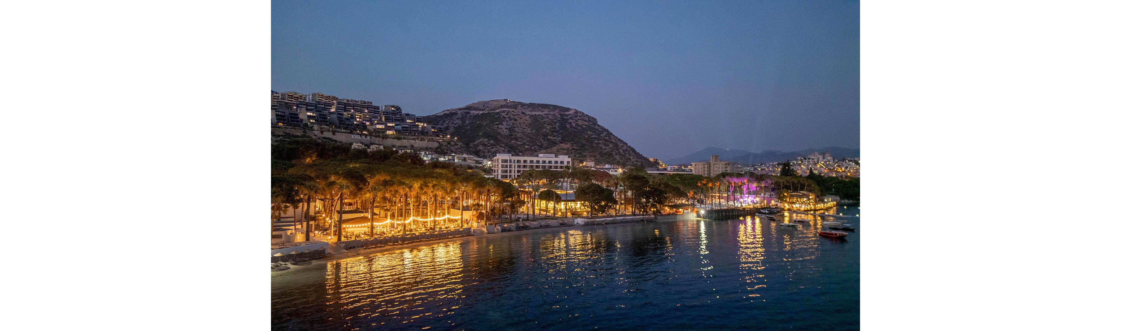 A coastal resort town is illuminated at dusk, with warm lights glowing among palm trees along the waterfront. Hillside apartments and hotels rise up the slope behind the shoreline, overlooking the calm sea. Reflections of the city lights shimmer on the water, while distant mountains fade into the evening sky. The overall atmosphere is tranquil and inviting, suggesting nightlife and leisure by the coast.