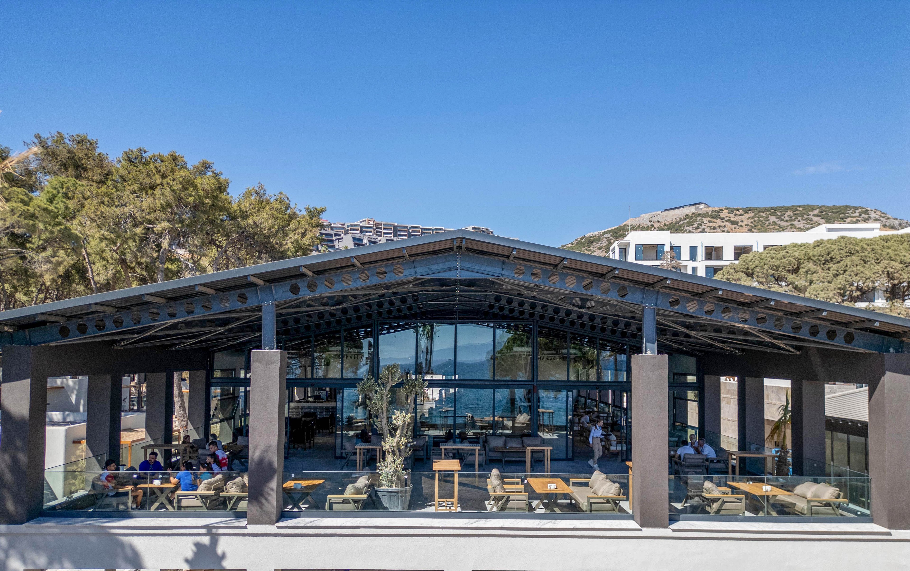 A contemporary glass-fronted restaurant with an open-air design is shown under a clear blue sky. The structure features a large peaked roof, dark columns, and expansive windows overlooking the sea. Outdoor seating with tables, chairs, and lounge areas is arranged on the terrace, where several people are dining and relaxing. Trees and hillside buildings frame the setting, creating a relaxed coastal atmosphere.