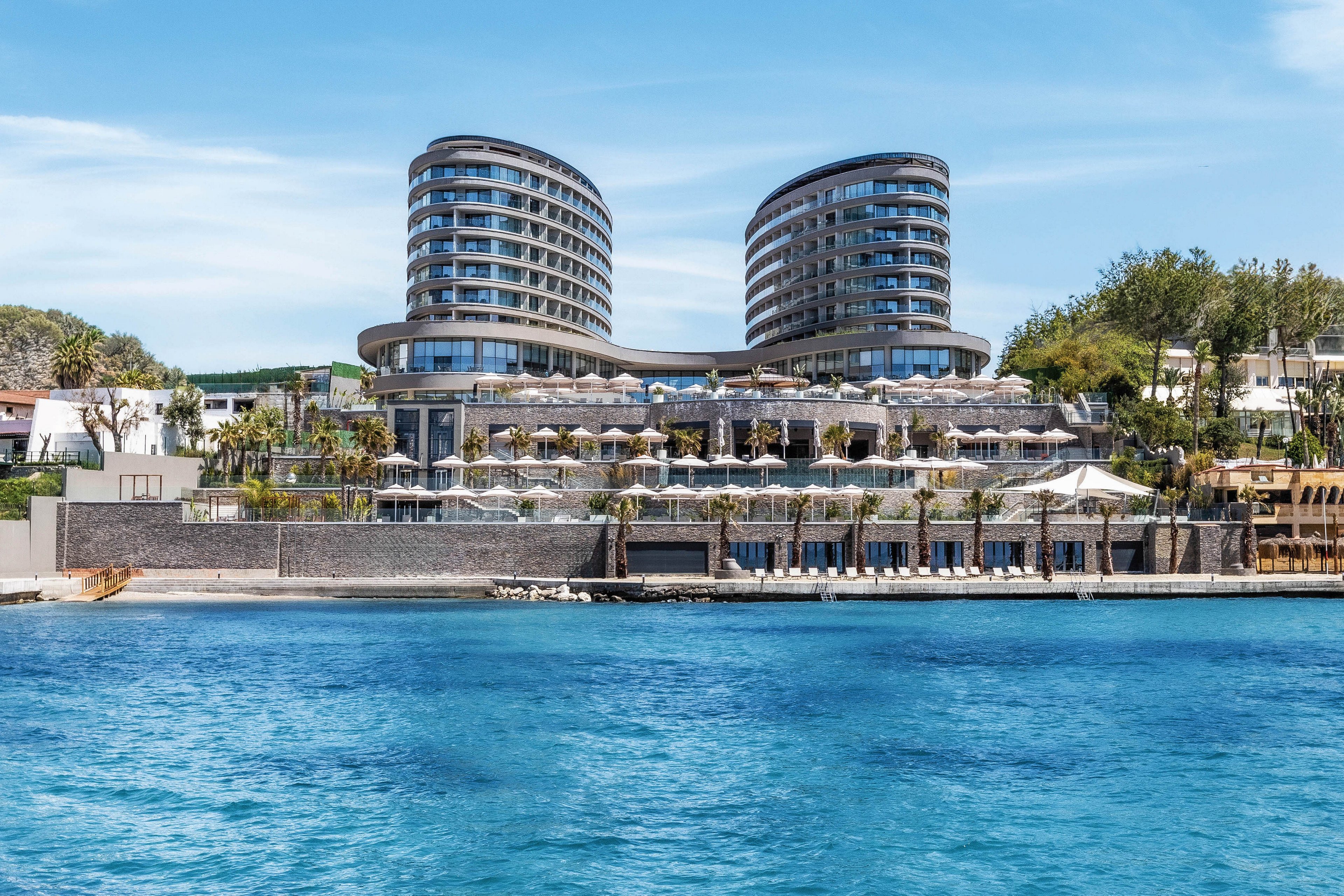 A contemporary seaside hotel with two curved twin towers rises above a calm blue waterfront. The building features multiple terraces, glass facades, and rows of sun umbrellas along the shore. Clear turquoise water fills the foreground under a bright blue sky with no visible clouds. Trees and smaller structures surround the main hotel complex, suggesting a resort destination.