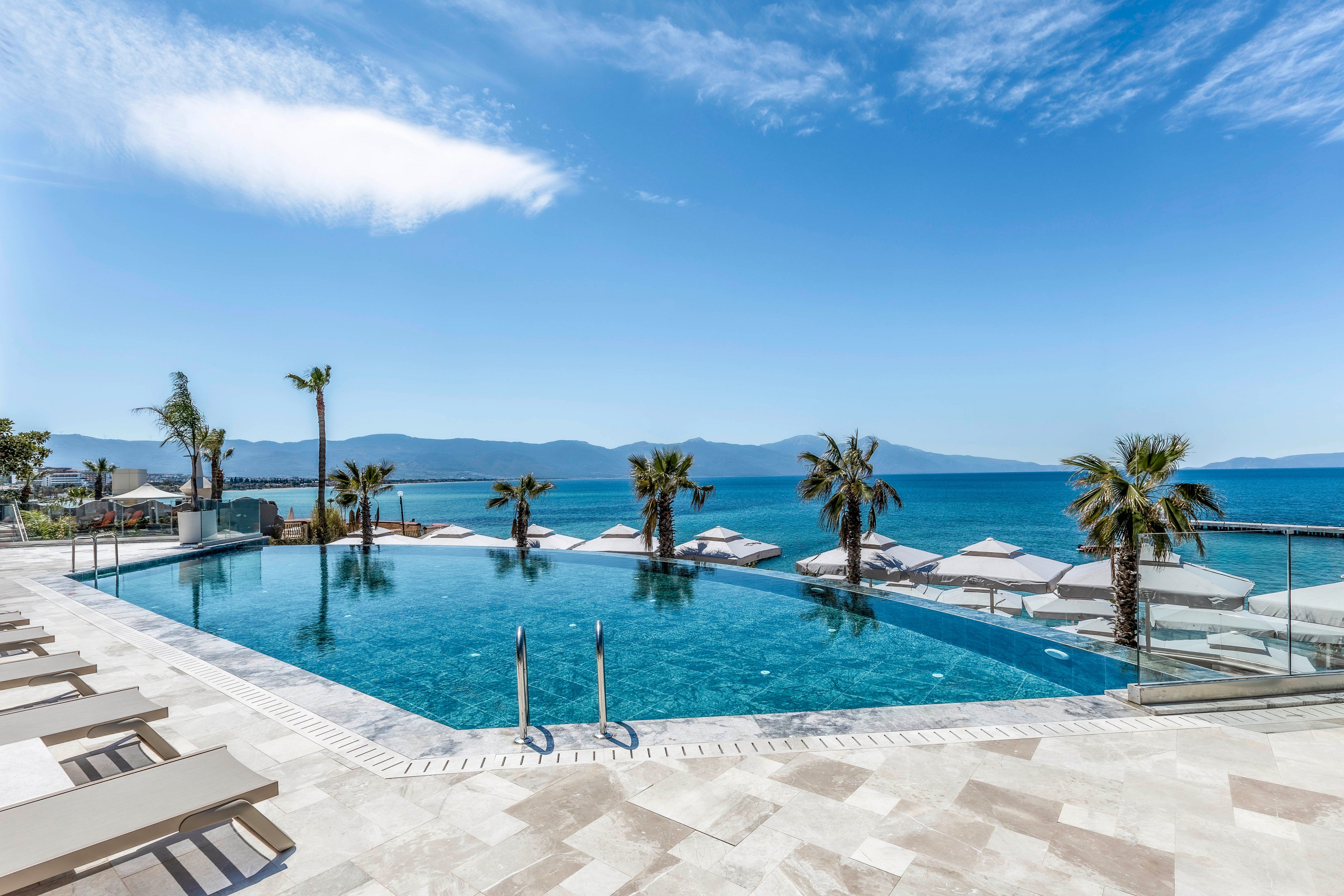 An elegant infinity pool stretches toward a calm blue sea under a clear sky. Sun loungers and umbrellas line the stone-tiled terrace, creating a relaxing resort atmosphere. Palm trees are placed around the pool, adding a tropical touch to the coastal setting. Distant mountains frame the horizon, enhancing the sense of openness and escape.
