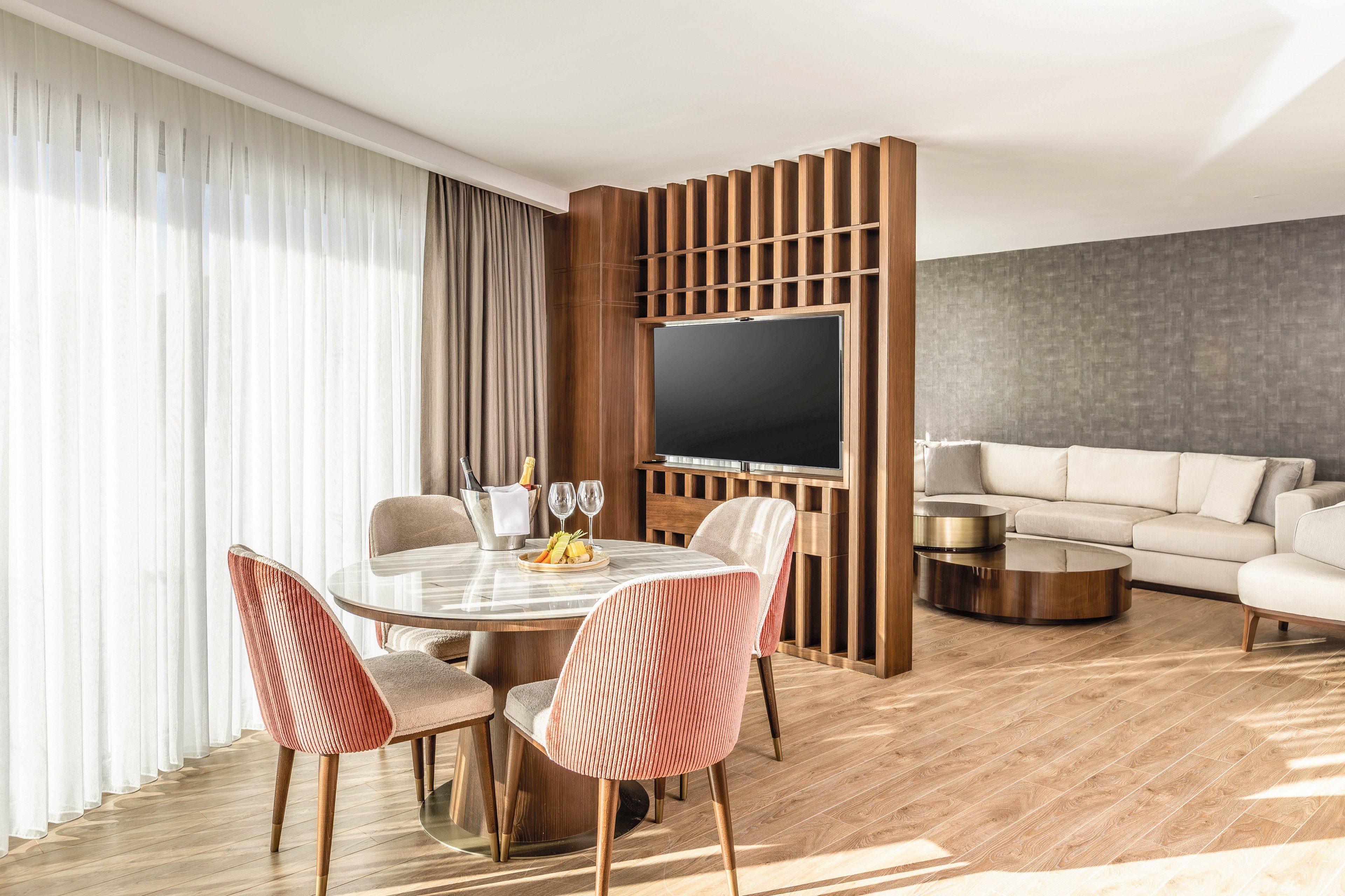 A contemporary hotel suite features a round dining table with four upholstered pink chairs in the foreground. Floor-to-ceiling sheer curtains allow abundant natural light to fill the space, highlighting the warm wood flooring. A wooden partition with built-in flat-screen television separates the dining area from a cozy seating area with a sectional sofa and round coffee table. The overall design is minimalist and elegant, with neutral tones and soft textures creating a relaxed atmosphere.