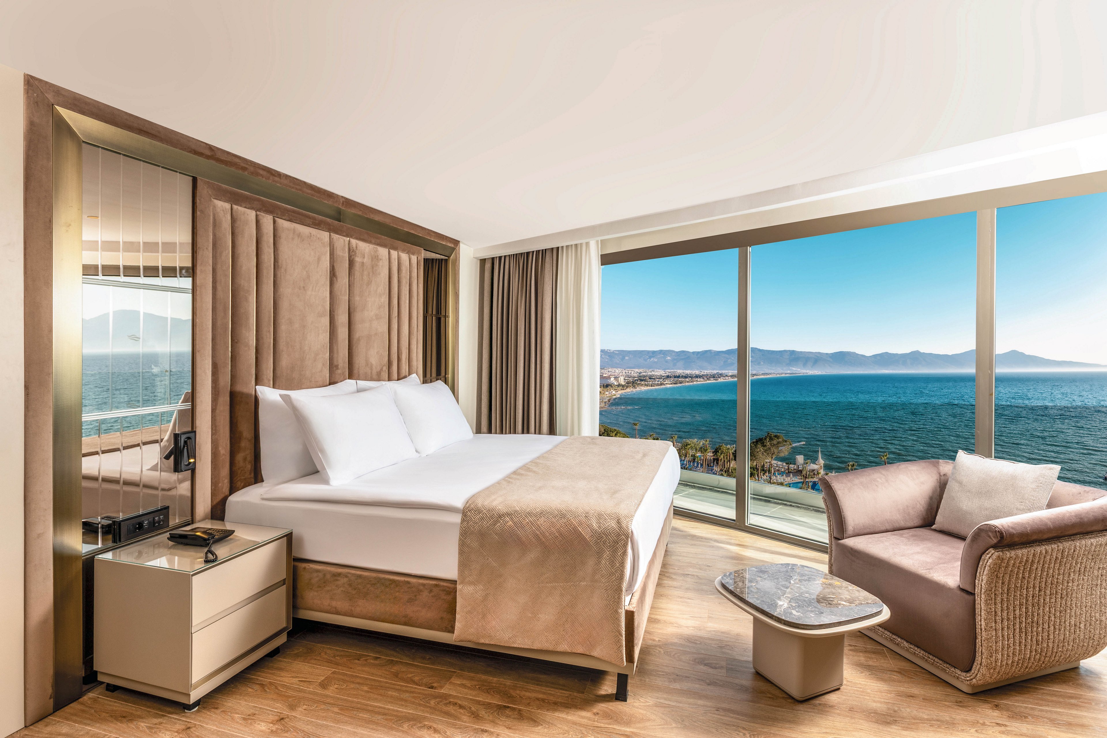 A contemporary hotel bedroom features a large neatly made bed with neutral-toned linens and padded headboard. Floor-to-ceiling windows reveal a sweeping view of the ocean, coastline, and distant mountains. A cozy armchair and small stone-topped table create a relaxing seating area beside the window. Warm wood flooring and soft beige and cream colors give the room a calm, luxurious atmosphere.