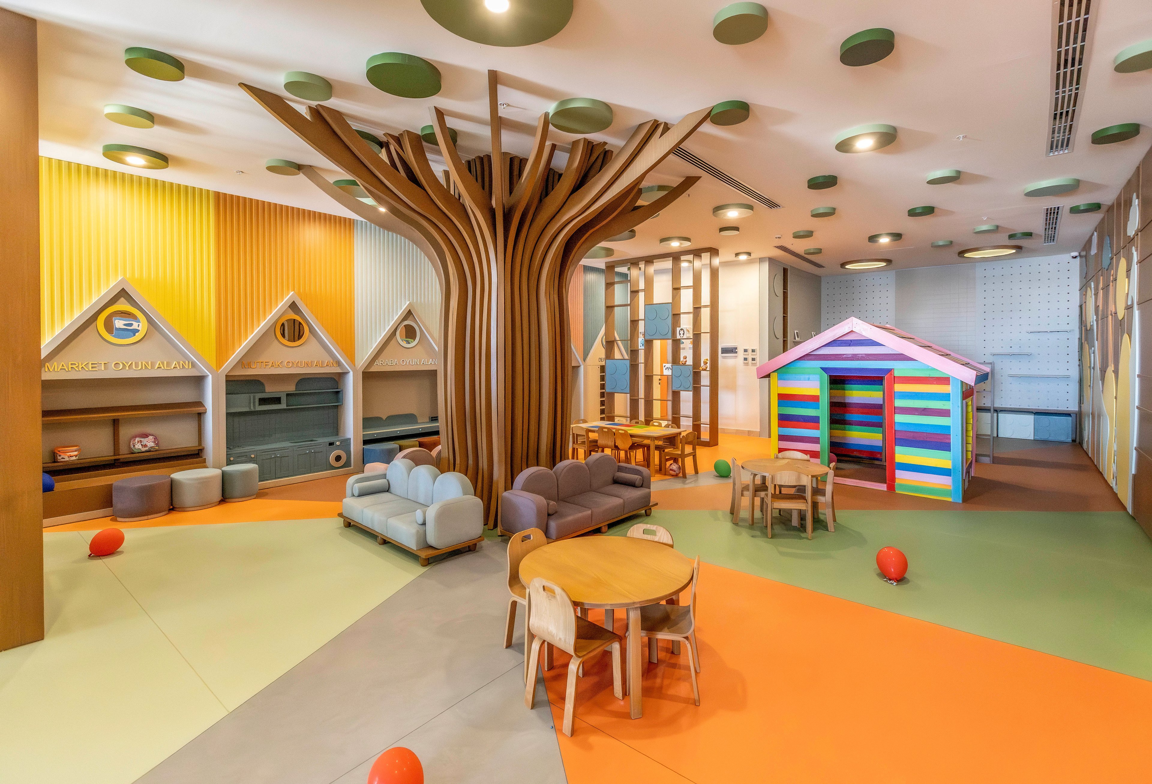 Spacious indoor children’s playroom featuring a large stylized tree structure in the center. The room includes small wooden tables and chairs, cushioned seating, and several playhouses, including a brightly colored striped house. Walls are decorated with house-shaped alcoves and warm pastel panels, while the floor is divided into orange, yellow, and neutral sections. Red balloons are scattered around the space, adding to the playful atmosphere. The overall design feels modern, safe, and inviting for young kids.