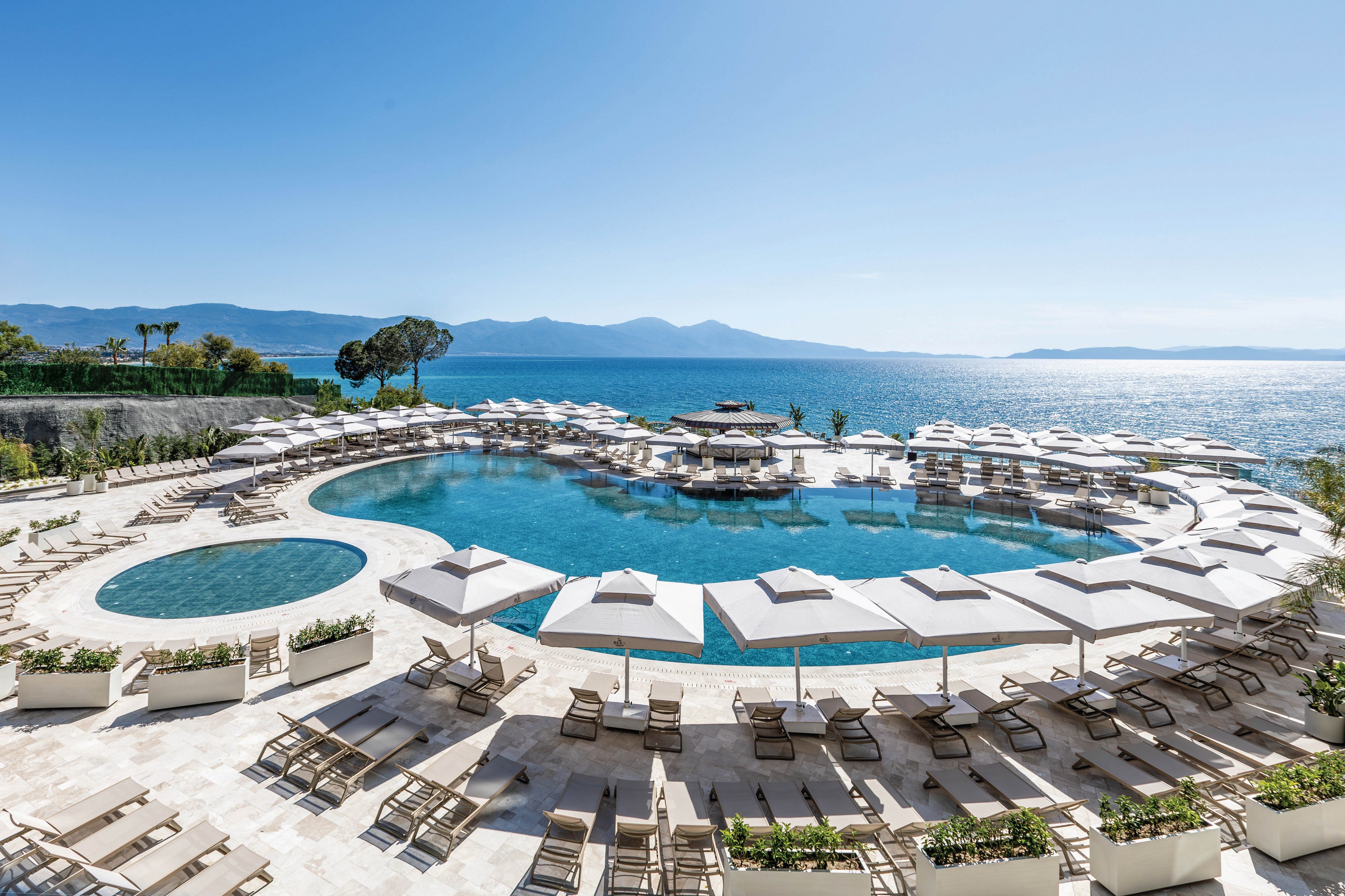 A large luxury resort pool complex overlooks a calm blue sea under a clear sky. Multiple circular and curved swimming pools are surrounded by rows of beige sun loungers and white umbrellas. The bright stone terrace contrasts with the deep blue water and distant mountains on the horizon. The scene is empty of people, emphasizing tranquility and a relaxing vacation atmosphere.