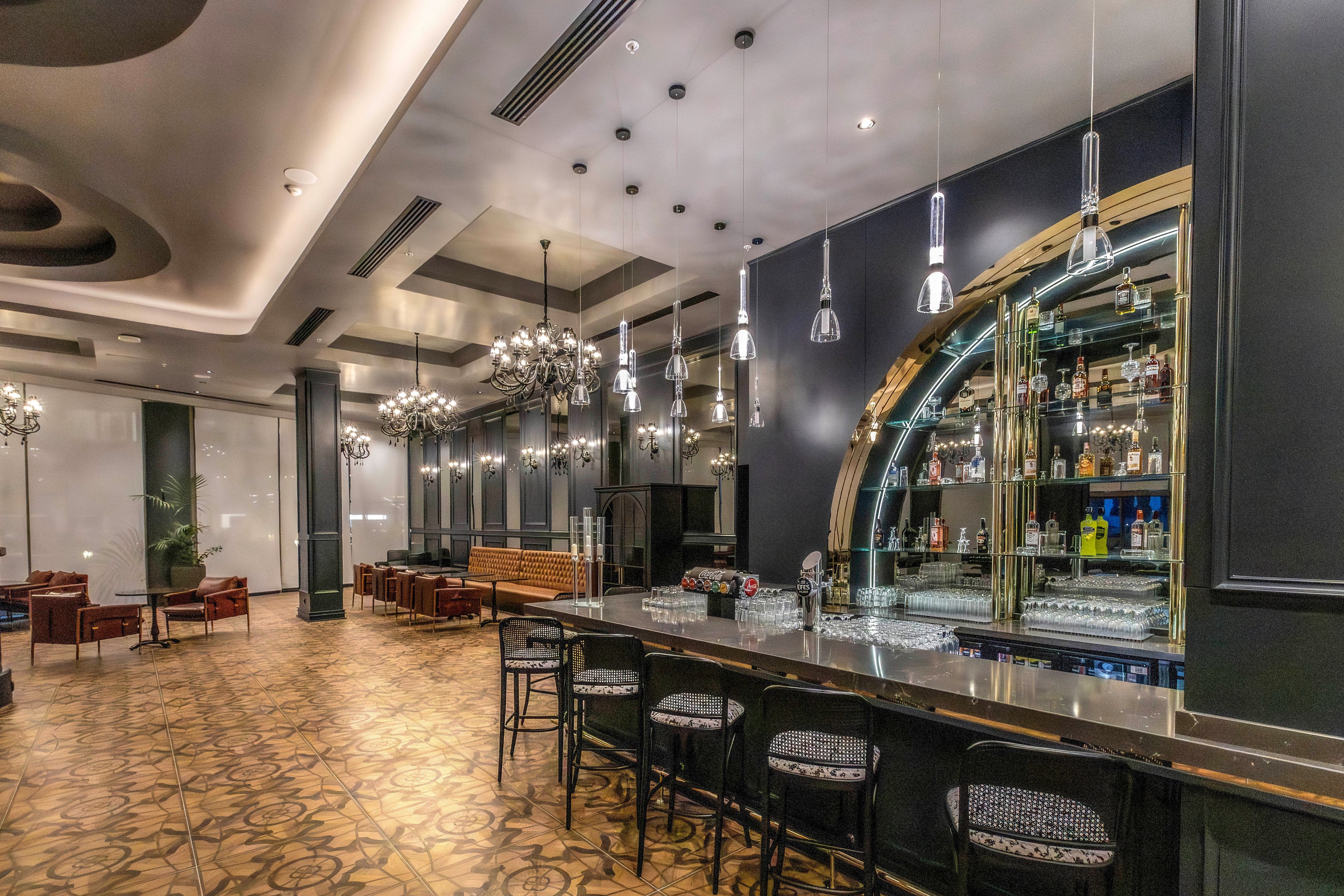 A contemporary hotel bar features a long stone counter with bar stools and a mirrored back bar stocked with bottles and glassware. The space opens into a lounge area with leather armchairs, wooden benches, and small tables arranged for conversation. Decorative chandeliers and pendant lights hang from a high ceiling, casting warm light across patterned tile flooring. Dark walls and metallic accents create a sophisticated, upscale atmosphere suitable for evening socializing.