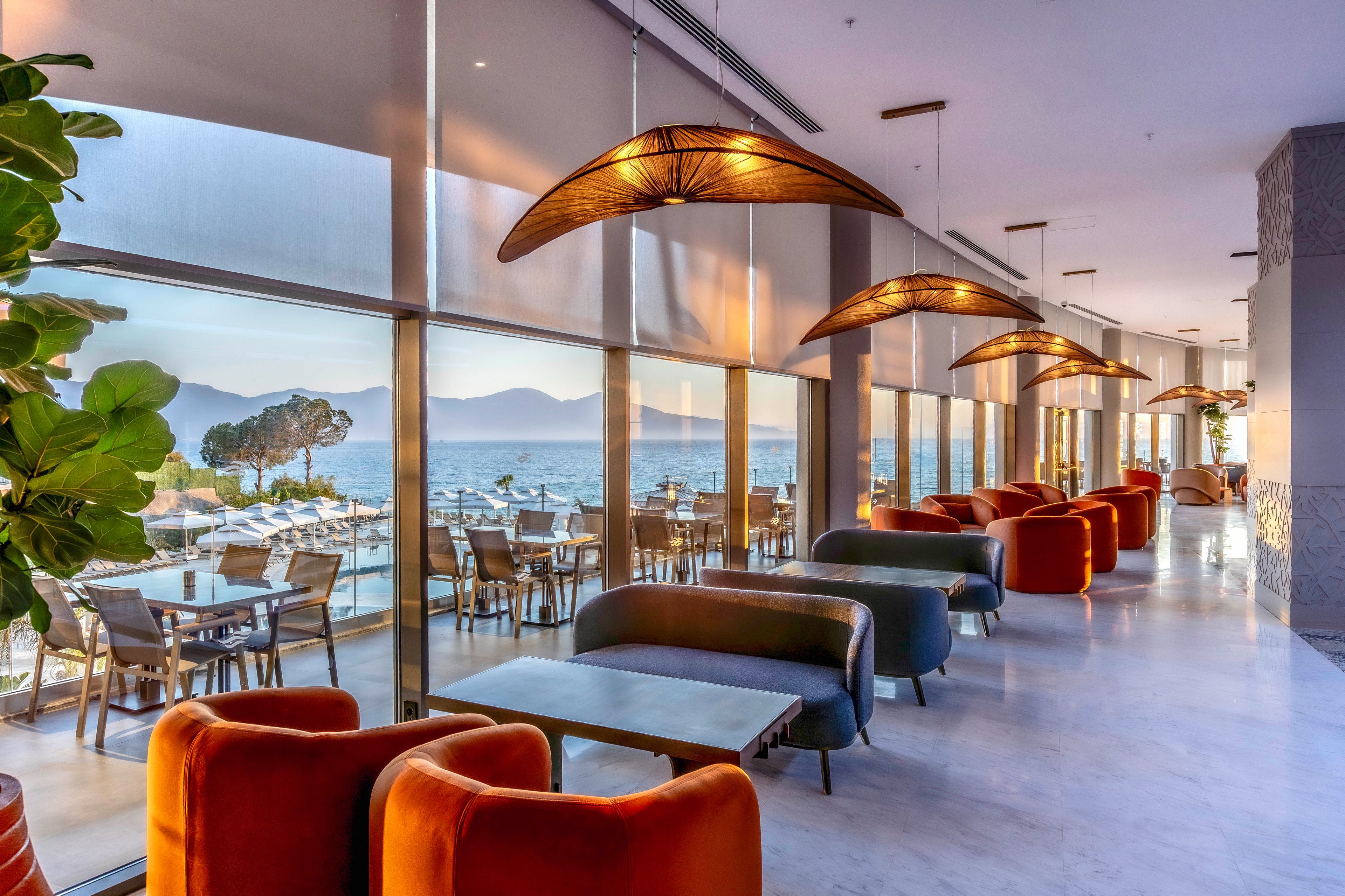 A contemporary hotel lounge features plush armchairs and low tables arranged along floor-to-ceiling windows. Outside, a terrace with dining tables overlooks a calm sea and distant mountains at sunset. Warm pendant lights and soft orange and brown seating contrast with the cool blue tones of the water and sky. The space feels relaxed and upscale, ideal for socializing or unwinding with a view.
