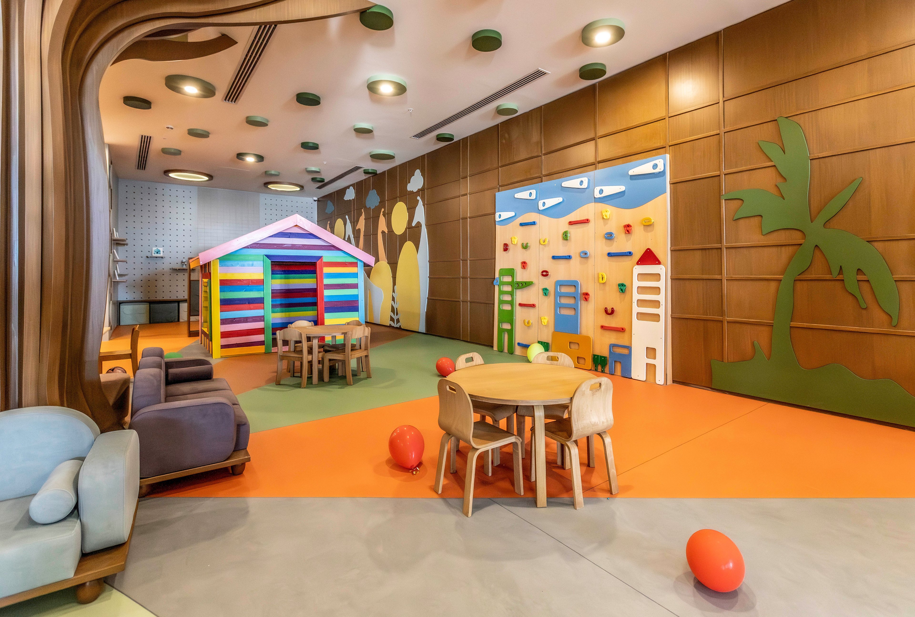 A bright indoor children's playroom features a colorful playhouse and small wooden tables with chairs. The space includes soft seating, scattered balloons, and a playful climbing wall with house-shaped elements. Warm wood paneling, whimsical wall art, and ceiling lights create a cheerful, inviting atmosphere. The room appears tidy and ready for kids to play and explore.