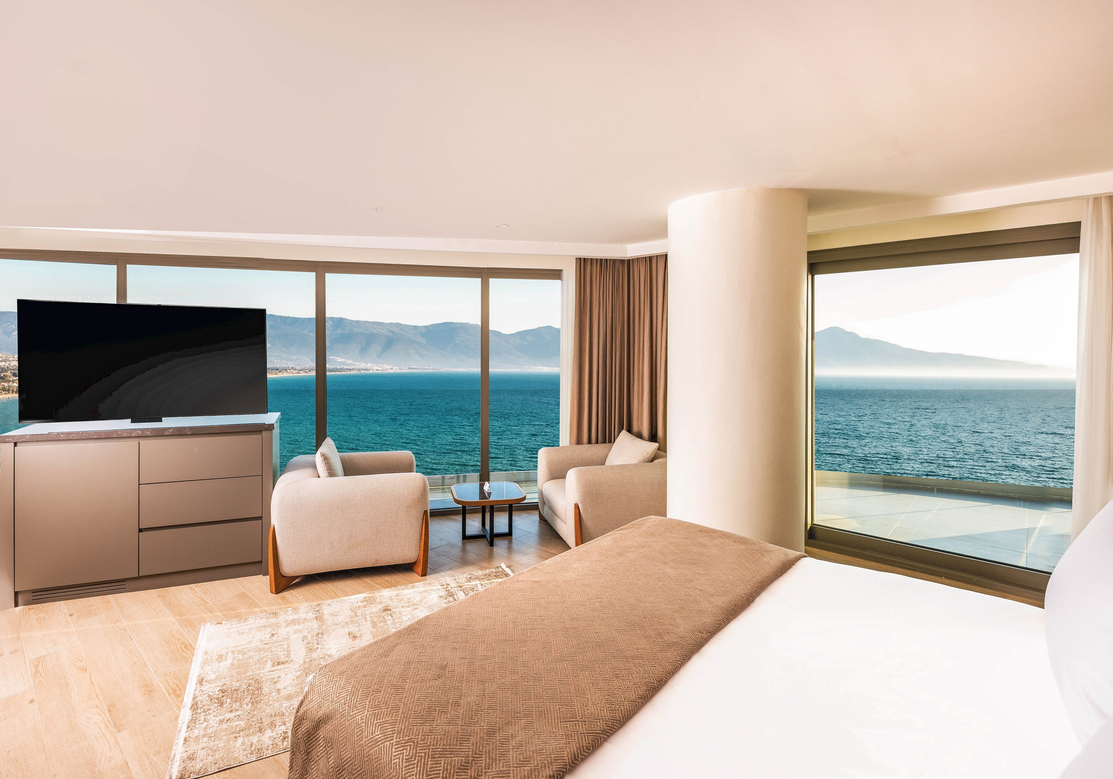 A contemporary hotel room features a large bed facing floor-to-ceiling windows with a sweeping ocean view. Two armchairs and a small table create a cozy seating area beside the windows. A flat-screen television sits on a sleek cabinet opposite the bed. Warm neutral tones, soft lighting, and minimalist decor give the space a calm, luxurious atmosphere.
