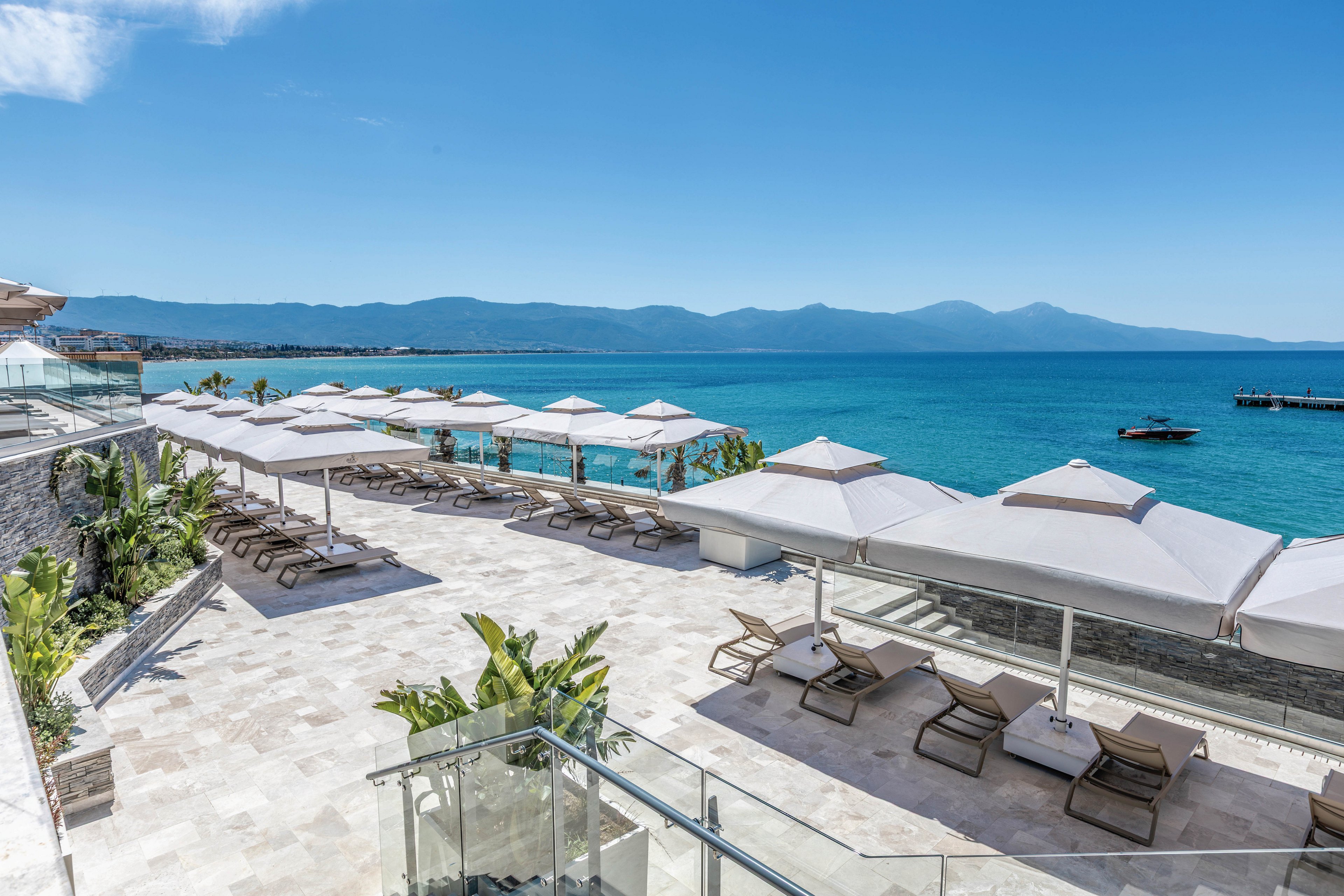 A spacious resort terrace overlooks a calm turquoise sea under a clear blue sky. Numerous sun loungers and large white umbrellas are neatly arranged on the light stone deck. Glass railings and tropical plants add a modern, upscale touch to the outdoor space. Distant mountains and a small pier with a boat complete the serene coastal setting.