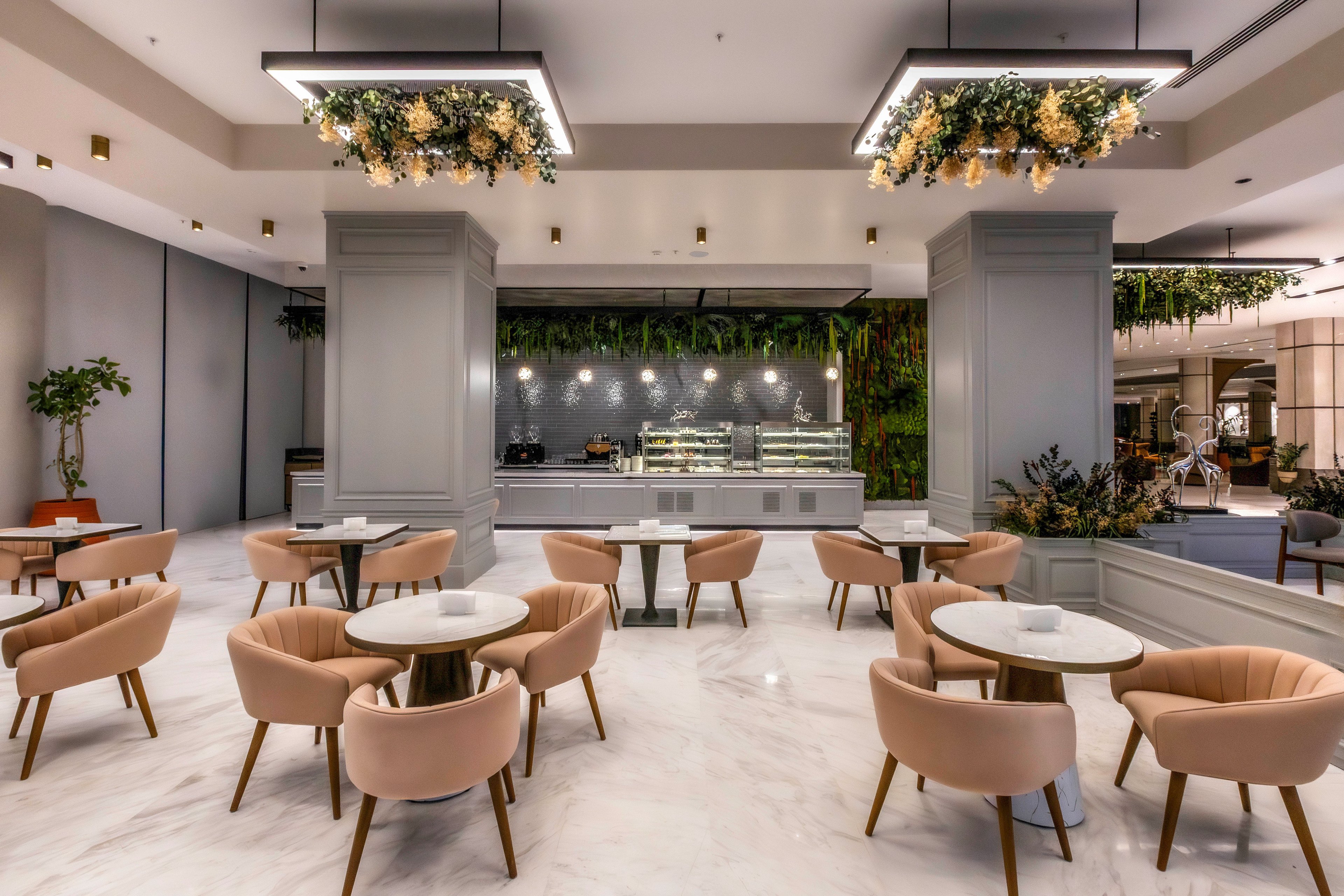 Contemporary café interior featuring round tables and plush upholstered chairs arranged on a polished marble floor. The space includes a central service counter with glass display cases and hanging greenery above. Warm pendant lights and decorative ceiling fixtures create a welcoming ambiance. Neutral tones of beige, gray, and white dominate the palette, accented by natural plants and subtle metallic details.