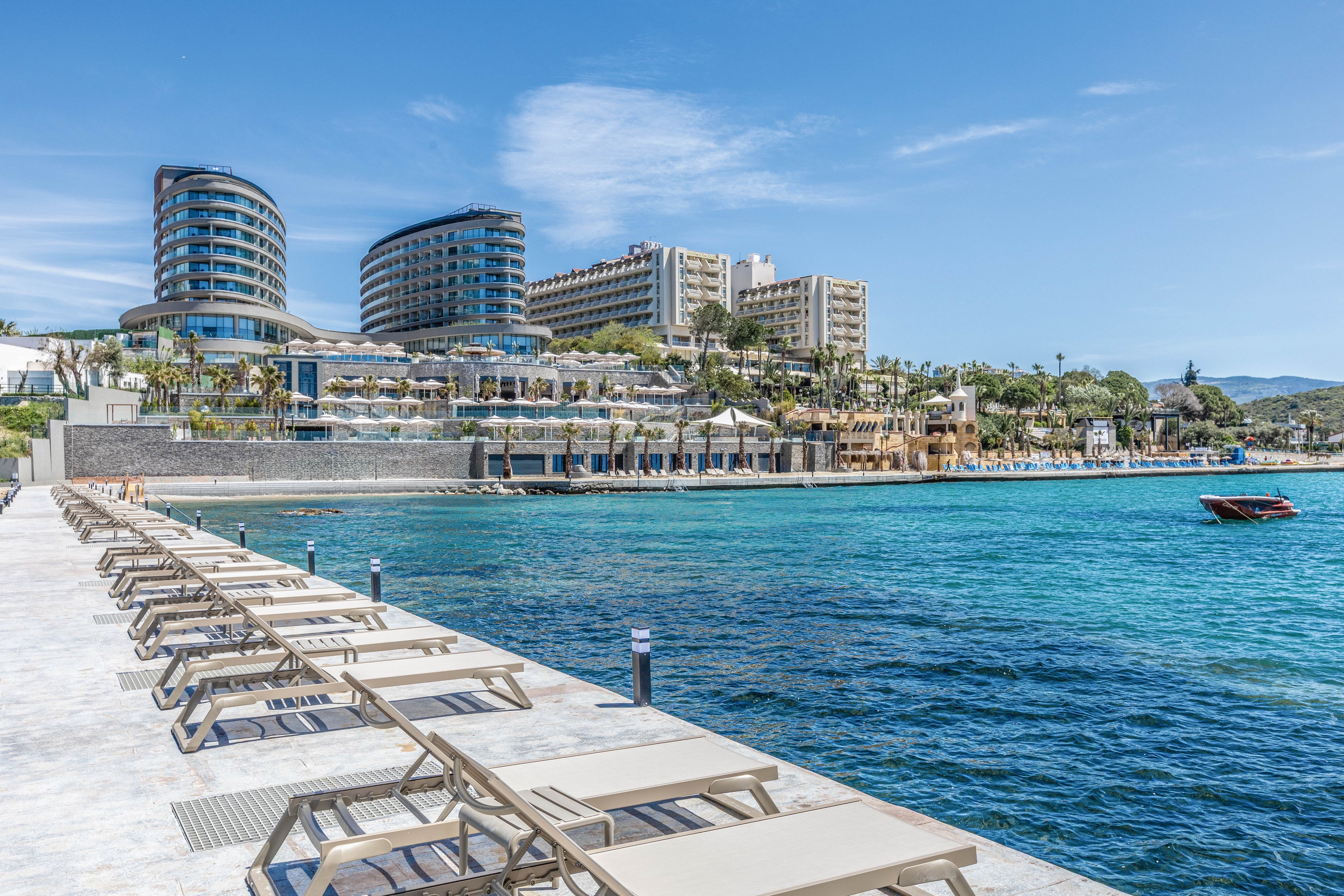 A row of empty sun loungers lines a bright waterfront promenade beside clear turquoise sea. In the background, modern curved high-rise hotels and resort buildings rise above terraced outdoor dining and pool areas. The sky is clear and blue, suggesting a warm, sunny day at a coastal vacation destination. A small red boat floats on the calm water, adding a pop of color to the tranquil scene.