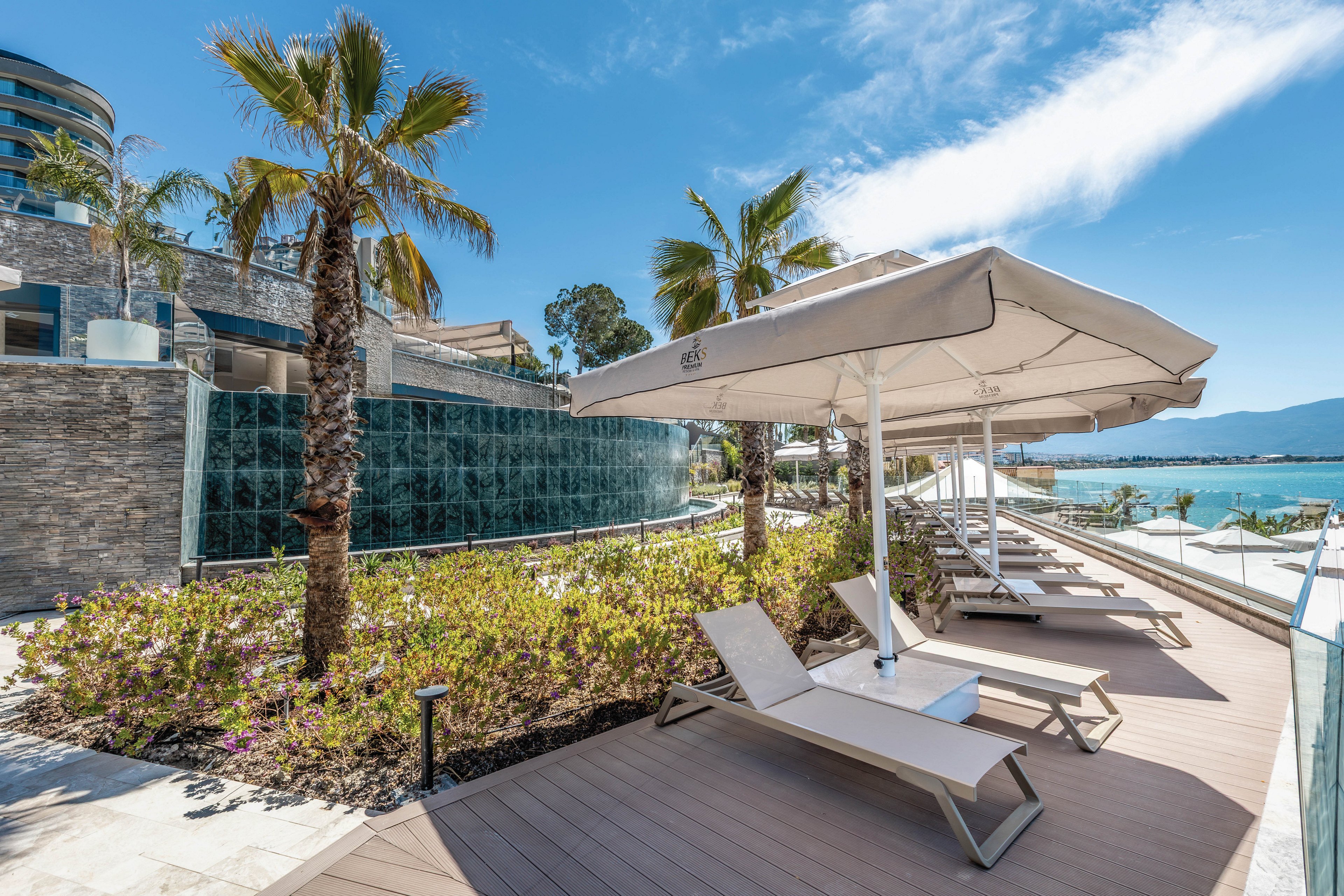 A modern seaside resort terrace features several empty sun loungers arranged under large white umbrellas. Palm trees and low flowering shrubs line the wooden deck, adding greenery to the contemporary stone and glass architecture. Beyond the terrace, a calm blue sea and distant mountains are visible under a clear, sunny sky. The scene suggests a peaceful vacation setting with ample space for relaxation by the water.