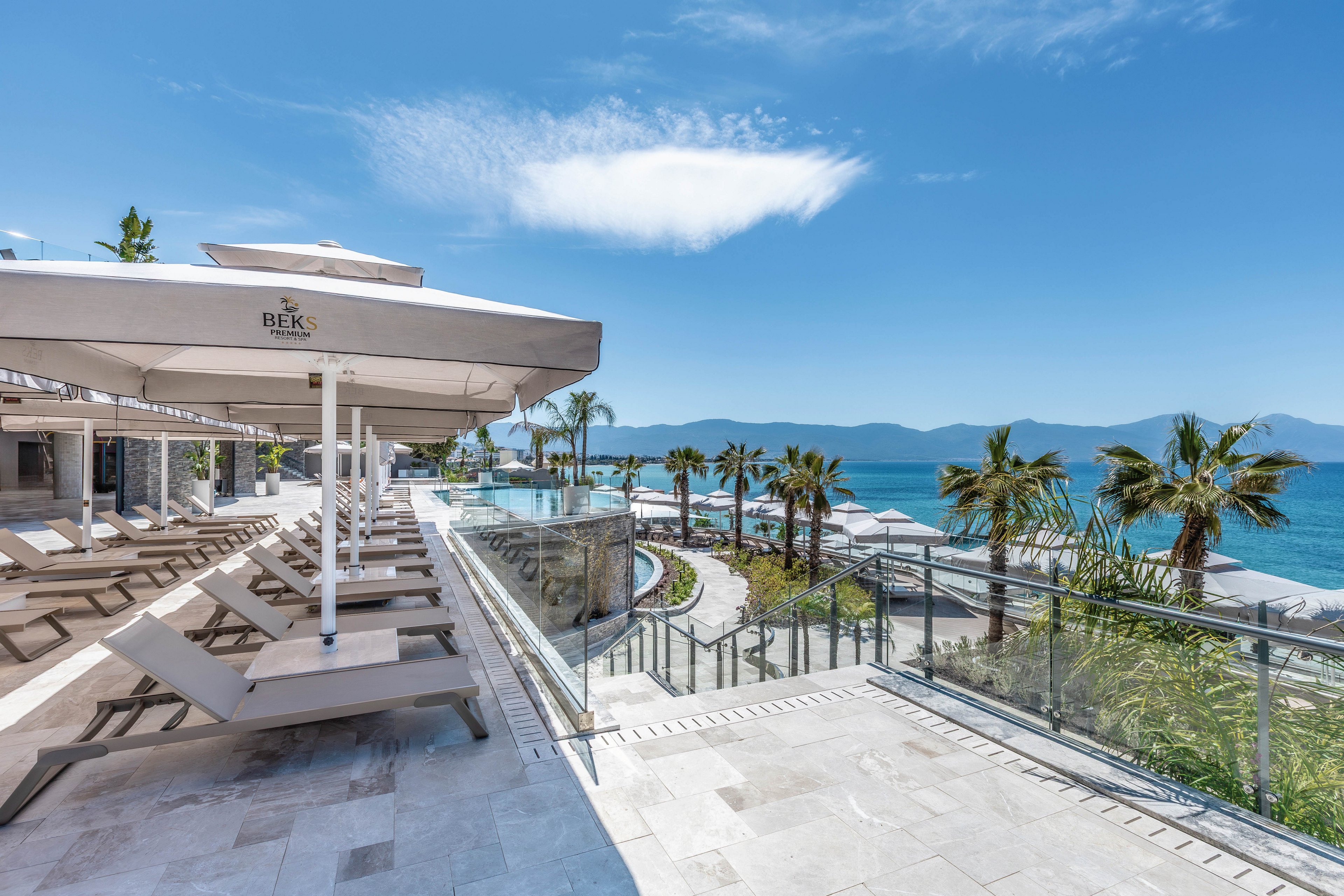 A modern resort terrace overlooks a bright blue sea and distant mountains. Rows of empty sun loungers sit beneath large white umbrellas on a clean stone deck. Glass railings frame the view of palm trees, landscaped paths, and the shoreline below. The clear sky and calm water create a serene, luxurious vacation atmosphere.